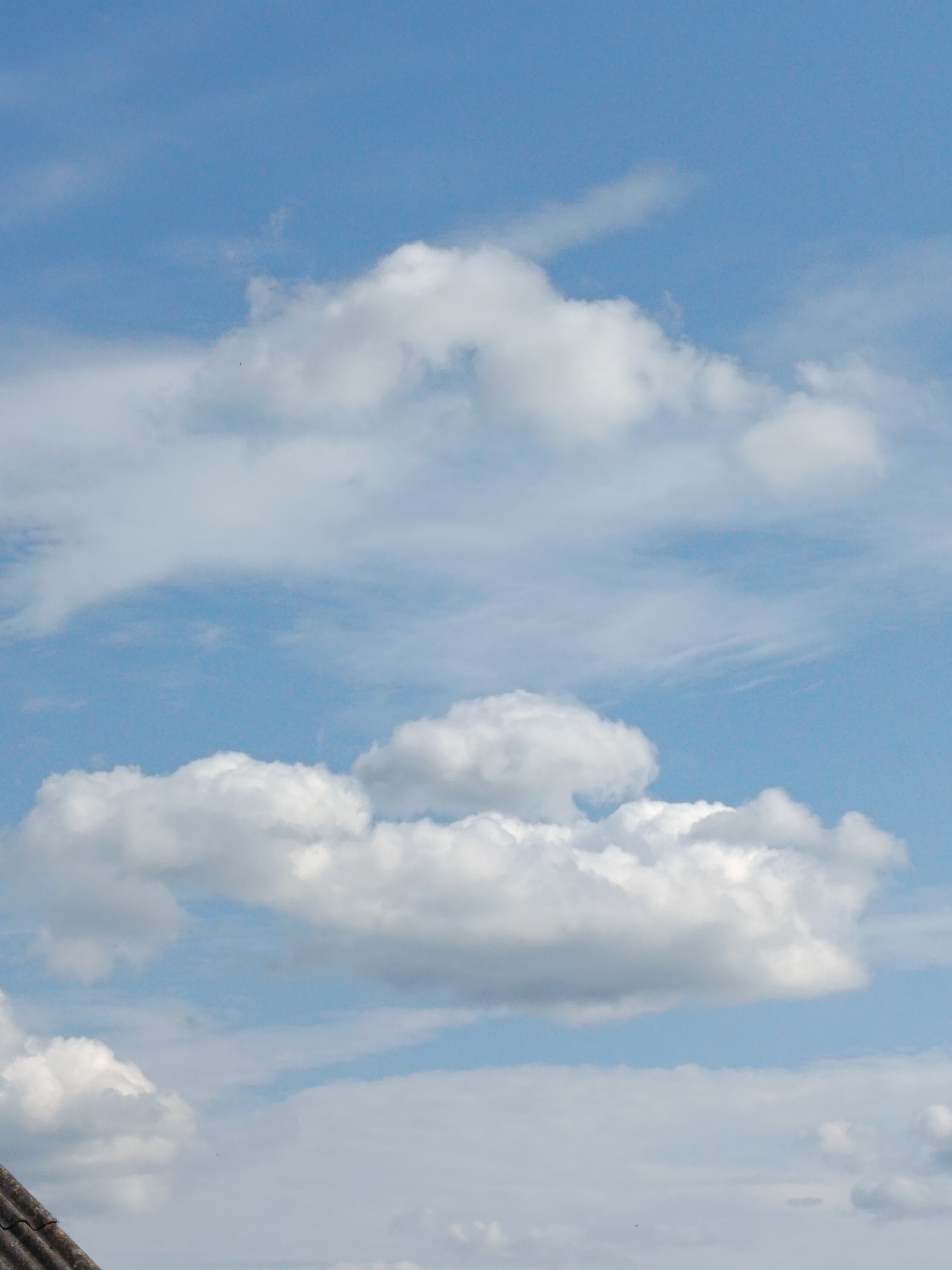 This is a photograph of a cloudy sky. The scene is set with a bright blue sky and scattered white clouds. The clouds are diverse in shape and size, with some appearing fluffy and cumulus-like, while others are more wispy and streaky. The bottom left of the image shows a small portion of a dark, corrugated rooftop, possibly indicating a building. The light suggests it is daytime, with the sun likely not directly visible but illuminating the clouds. There are no people or specific activities depicted. The overall impression is a peaceful, open sky, typical of a partly cloudy day. The location context provided is Novozybkov, Russia.