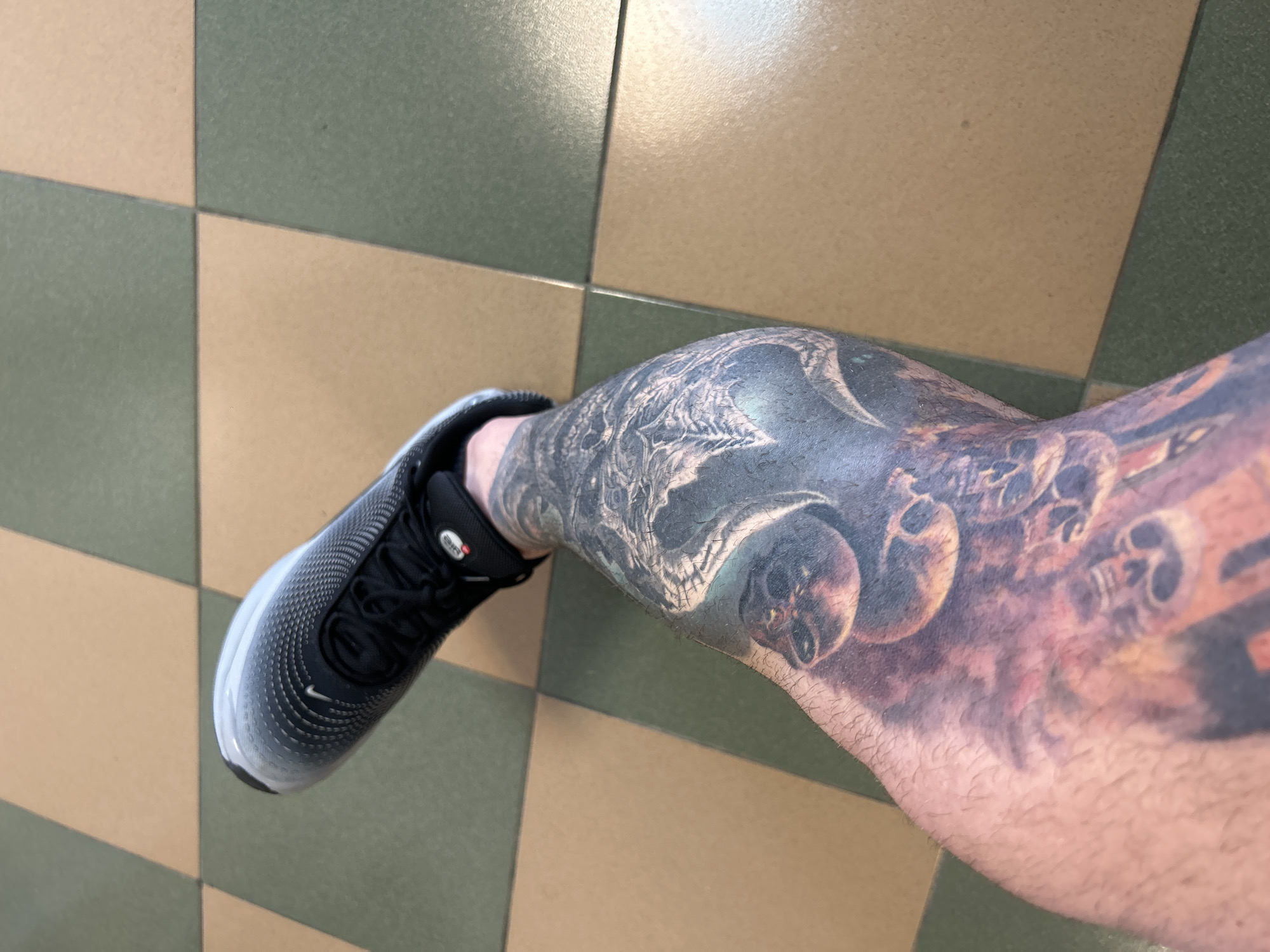 The image shows a lower left leg and foot wearing a black and gray Nike running shoe. The leg is extensively tattooed with a dark, detailed design featuring skulls and other gothic imagery. The background consists of a checkered floor of alternating beige and green tiles.
