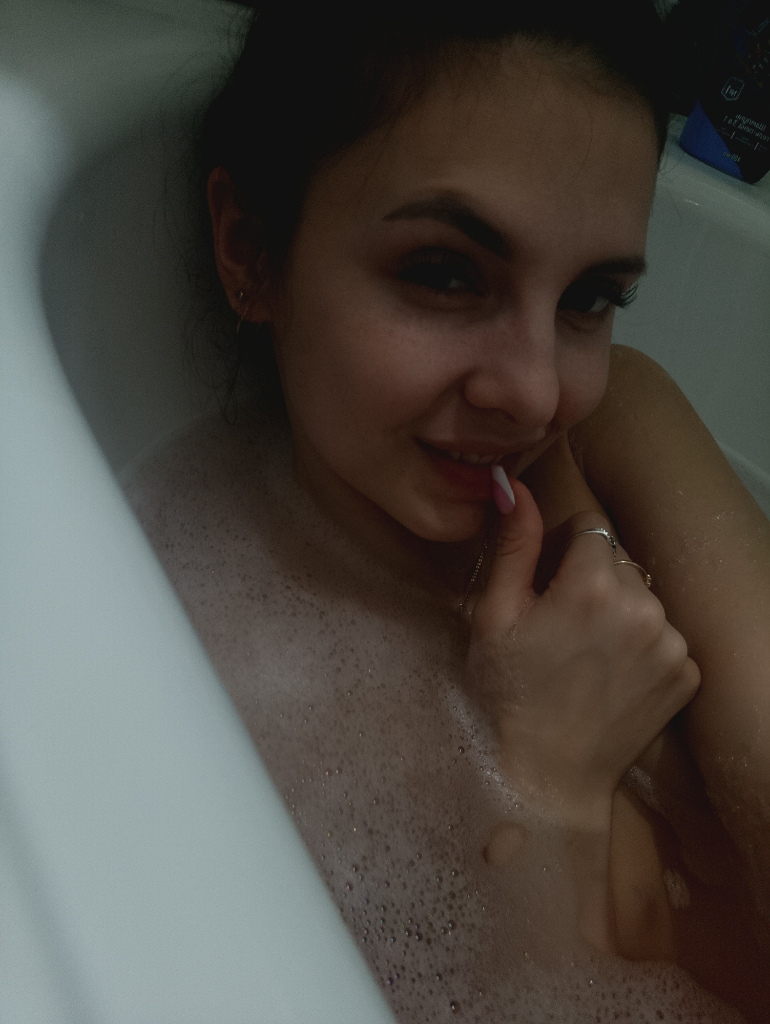 A young woman is partially submerged in a bubble-filled bathtub. Her face is visible, with dark hair pulled back, and she is smiling with her hand near her mouth. She is wearing rings on her fingers. A blue container is partially visible in the upper right corner of the frame. The location appears to be a bathroom in an unknown city.