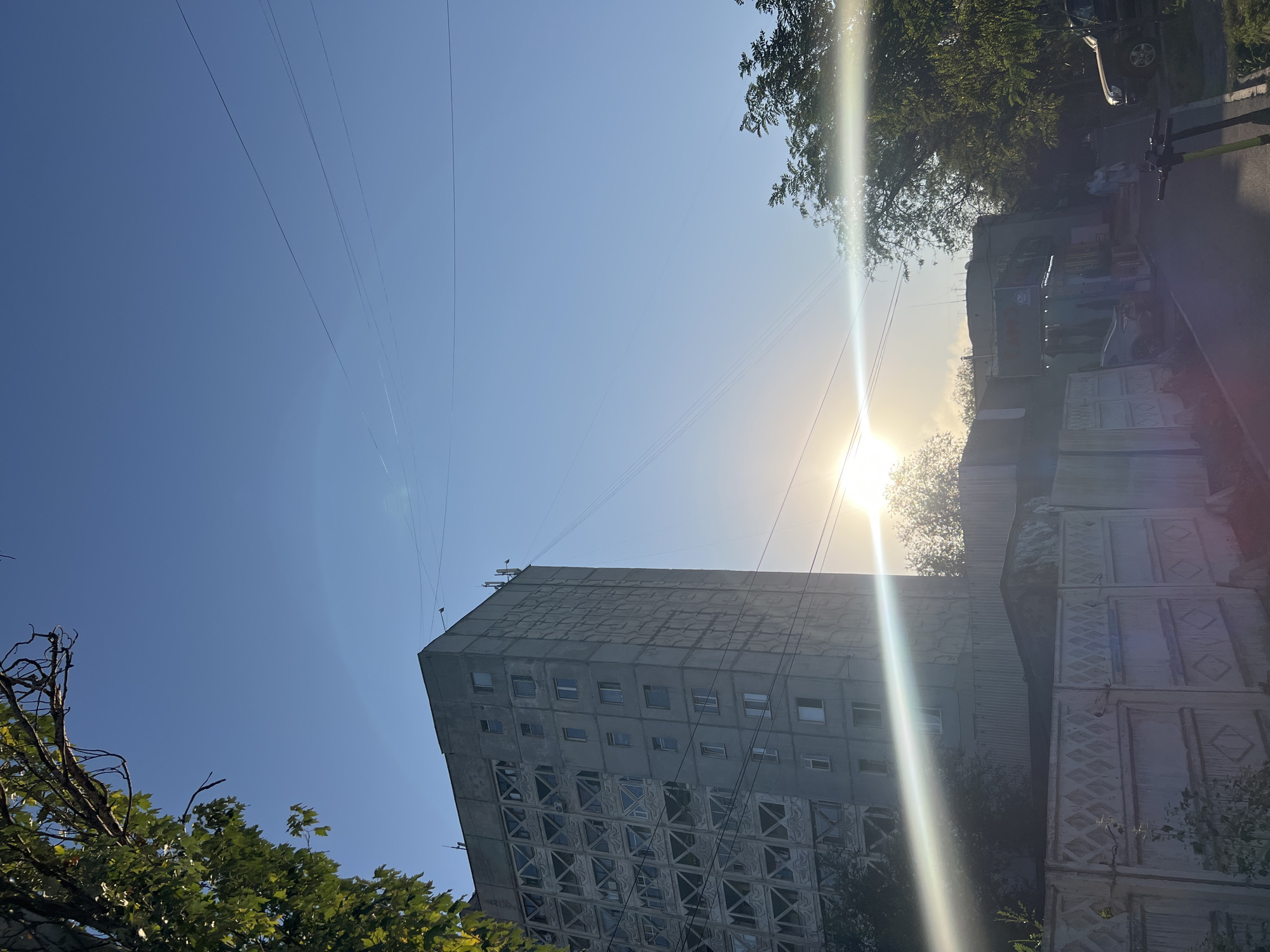 The image depicts a street scene in Bishkek, Kyrgyzstan, under a clear blue sky with the sun shining brightly, creating lens flares. In the foreground, a section of a patterned concrete fence runs horizontally. To the left of the fence, a tall, grey apartment building with a distinct geometric facade is visible. Trees with green foliage partially obscure the lower portion of the building and the area behind the fence. Further down the street, there are smaller buildings, some appearing to be shops, and more trees. Several power lines crisscross the sky above the buildings. On the street, there is a parked black SUV on the right and two electric scooters in the lower right corner. A few indistinct figures are walking further down the street. The overall impression is a residential or mixed-use urban environment on a sunny day.
