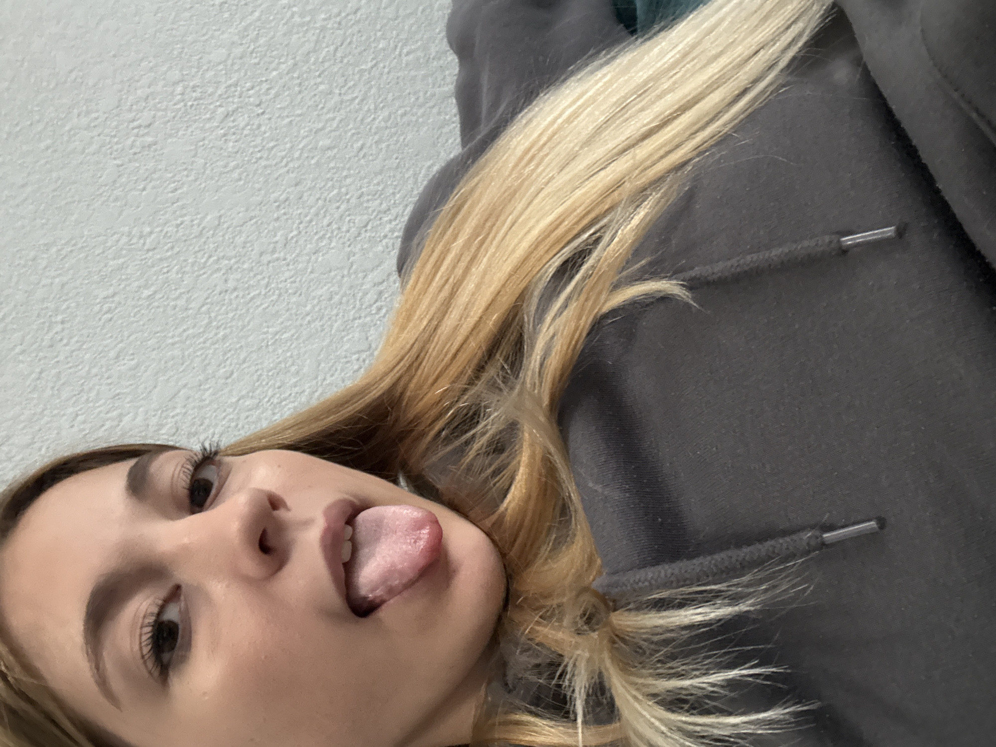 This image, set in Sofia, Bulgaria, captures a close-up portrait of a young woman from a tilted perspective. The primary subject is the woman, whose face and upper torso are visible, angled from the lower left towards the upper right. She has long, straight, light-colored (blonde or light brown) hair that cascades over her shoulder and arm. Her dark eyes are open, looking slightly to the right, framed by prominent eyelashes. She has dark eyebrows and a fair complexion.
The woman is engaged in a playful activity, sticking her tongue out. Her mouth is open, revealing a portion of her tongue, which appears pink and textured. She is wearing a dark grey or charcoal-colored hooded sweatshirt, a portion of which is visible covering her body. A black drawstring with a silver-colored metal aglet is visible on the hoodie.
The background consists of a plain, light-colored wall with a rough, textured finish, suggesting it could be stucco or painted concrete. The lighting appears to be indoor. No text is visible in the image. The overall impression is a casual, lighthearted moment captured in an informal setting.