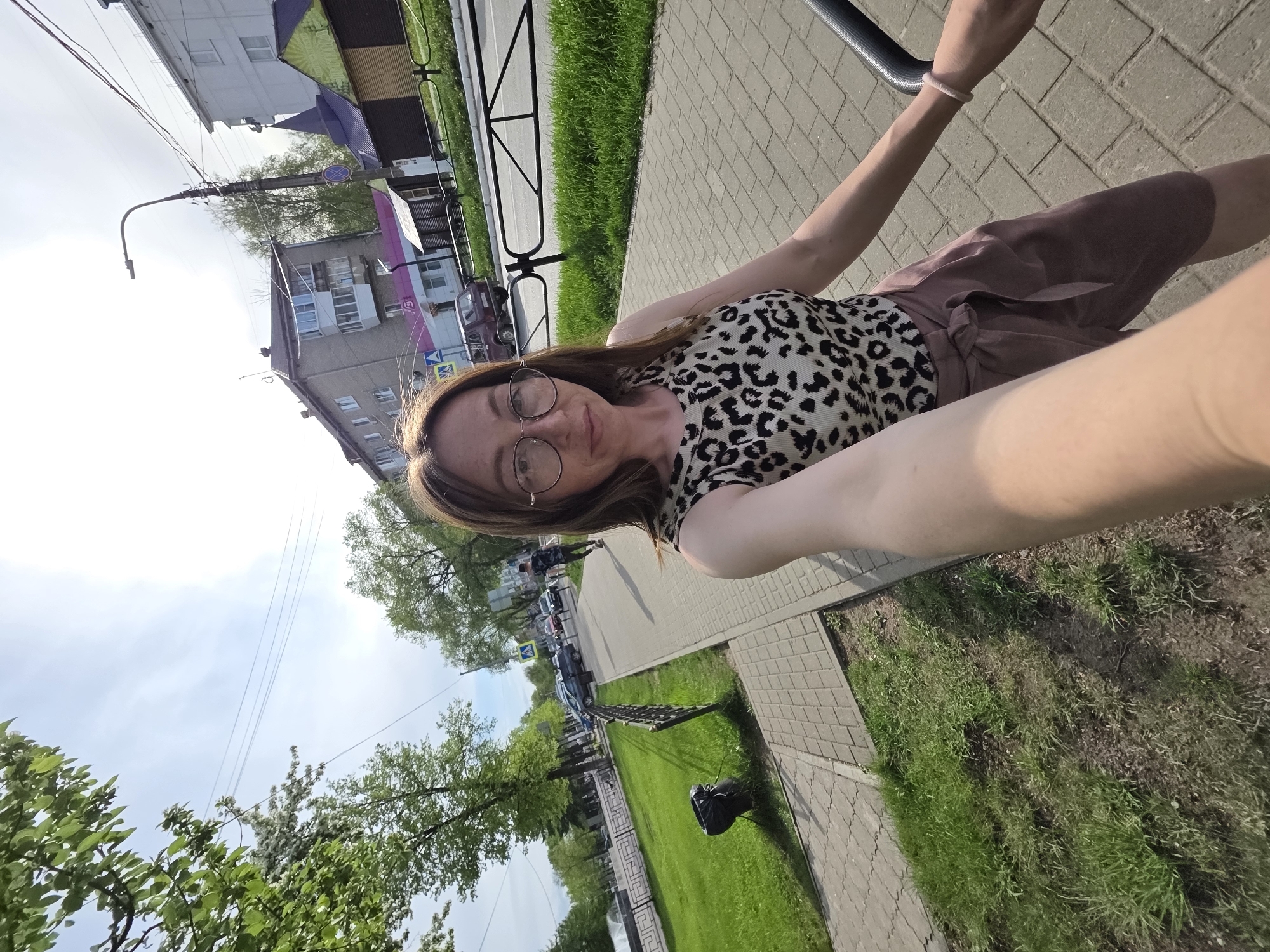 The image appears to be a selfie taken on a sidewalk in Chaikovsky, Russia. A woman with glasses and brown hair is in the foreground, wearing a leopard-print top and brown shorts. The sidewalk is made of grey paving stones, adjacent to a patch of green grass. In the background, there are buildings, trees, and parked cars along the street. There is a street light.