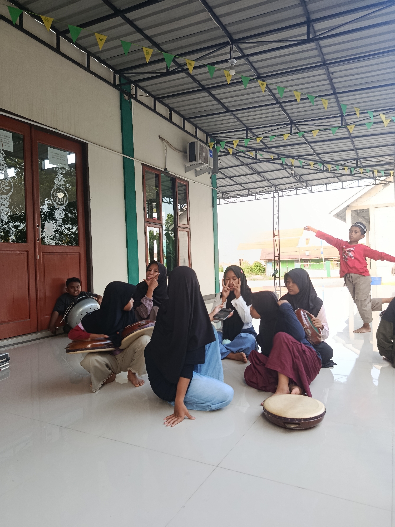 In Swarga Bara, Indonesia, a group of children and young women are gathered outdoors under a covered patio. The patio has a corrugated metal roof supported by a dark metal frame, and decorated with green and yellow pennant banners. A wall with two large wooden doors and windows, bearing Islamic calligraphy, forms the background on the left. The group is seated on a tiled floor.

In the foreground, a young boy in a red shirt and kufi stands, his arms outstretched. To his left, several young women, some wearing hijabs, are seated on the floor. One young woman in a black hijab is holding a phone. Another, wearing a dark purple skirt and black hijab, is holding a tambourine. Next to her, a young woman in a blue skirt and black hijab is also holding a tambourine.

To the left, a boy in a dark shirt sits near a large silver drum, leaning against the wall. A woman in a black hijab is seated with her back to the camera, facing the group. The scene appears to be a casual gathering, possibly involving music or a religious activity, given the presence of musical instruments and the Islamic decorations. The weather seems fair, with bright daylight suggesting it is daytime.