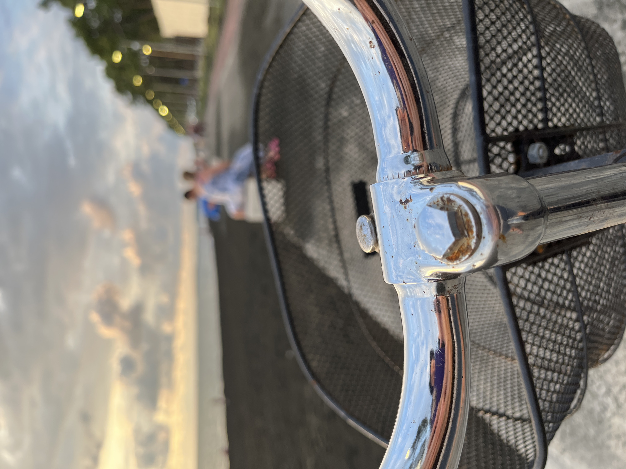 The image is a first-person perspective of a bicycle's handlebars and front basket. The handlebars are chrome and show signs of rust. Behind the handlebars, a black wire mesh basket is visible. In the background, a beach with dark sand meets a body of water. Further in the distance, a walkway lined with trees and streetlights is present, with a few blurred figures seated on a low wall. The sky above is cloudy with warm tones, suggesting it is either sunrise or sunset. The overall scene appears to be along the coast of Ureki, Georgia, a town known for its magnetic black sand beaches and therapeutic properties.
