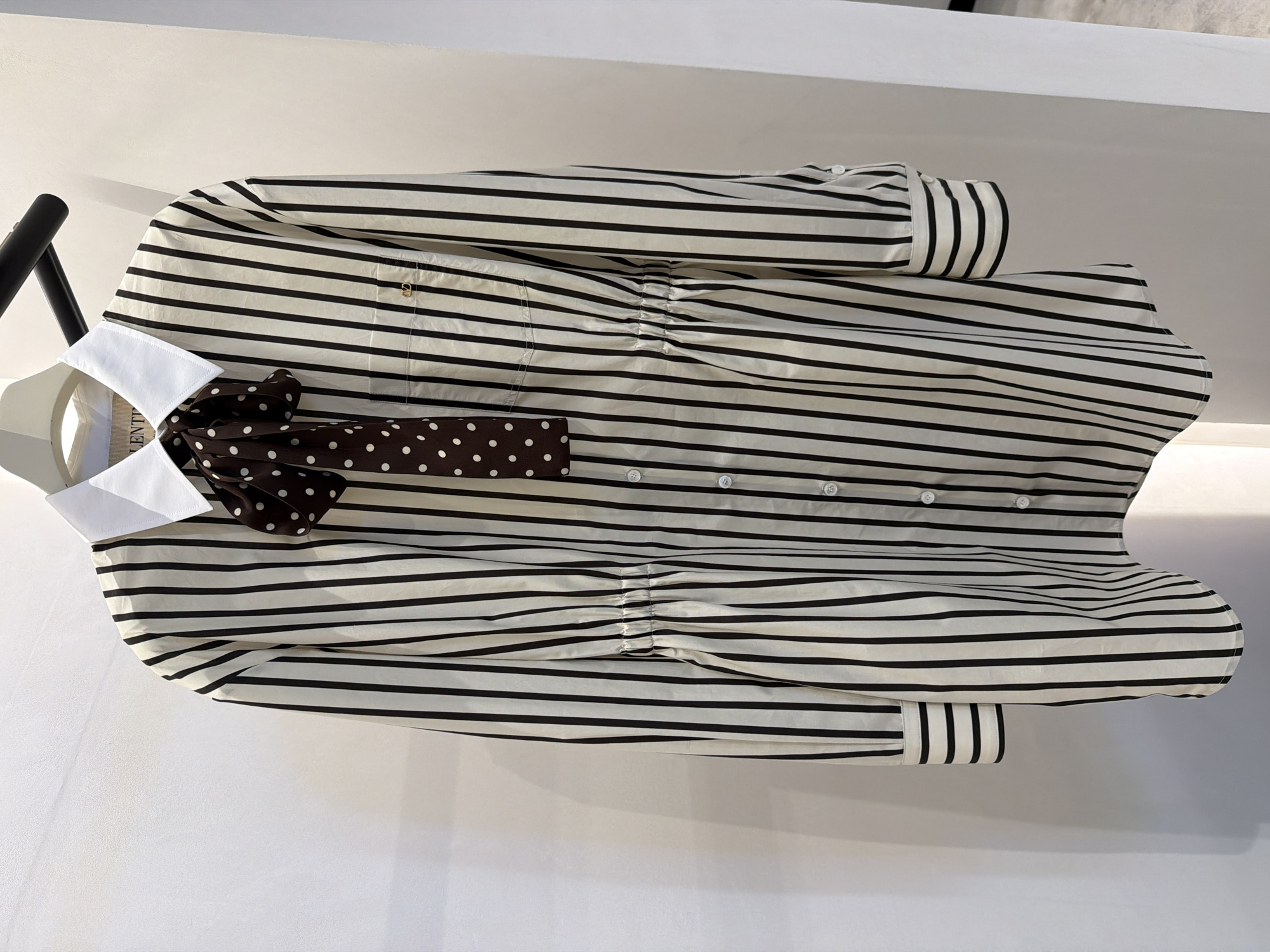 The image shows a vertical, full-body shot of a striped shirt dress on a white hanger against a white wall. The dress has a white collar and a brown polka-dot bow tie attached. It features vertical black and white stripes, a button-down front, a chest pocket with a small gold logo, and a cinched waist with elastic gathering. The sleeves have buttoned cuffs with horizontal stripes. The background is a plain white wall with some light and shadow. The image is taken from a medium distance, allowing for a clear view of the garment's details. There is no indication of the location being in New York City, United States of America.