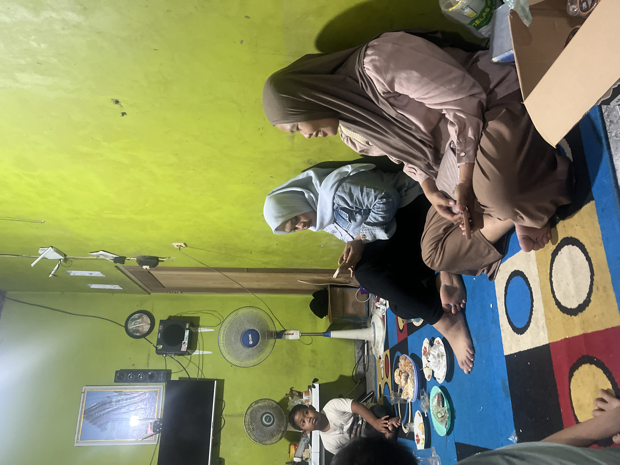 The image depicts an indoor gathering likely in a residential setting in Wedarijaksa, Indonesia. Two women, both wearing hijabs, are seated on a colorful rug on the floor. The woman on the right is wearing a pink top and brown hijab, and is smiling while looking to her right. The woman on the left is wearing a denim jacket over a white shirt and a light blue hijab, and is looking down at her phone. In the foreground, plates of food and drinks are laid out on the rug. To the left, a young boy in a white t-shirt is sitting on the floor, looking to his right. In the background, a wall painted a bright lime green dominates the scene. Various objects are visible on the wall, including a framed picture, speakers, a clock, and a ceiling fan. Two standing fans are also present in the room. The lighting appears to be artificial, creating shadows on the wall. The overall atmosphere suggests a casual social gathering.