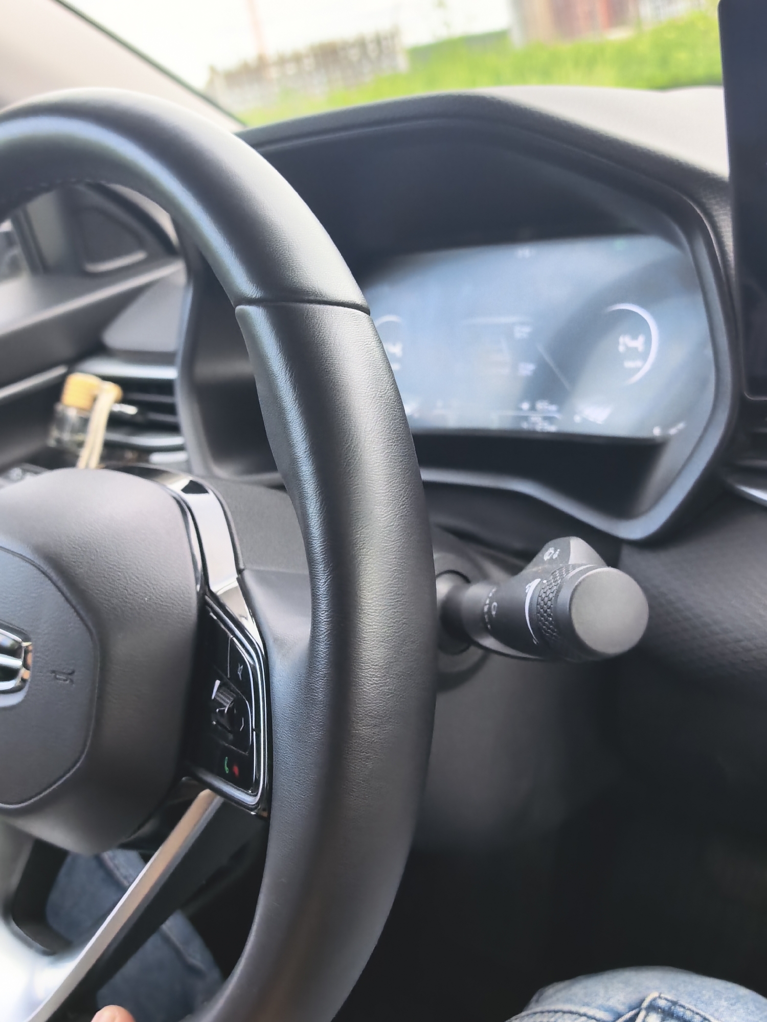 A close-up shot shows the interior of a car, focusing on the steering wheel and dashboard. The steering wheel is black leather, with chrome accents and buttons on the right side. To the right of the steering wheel is a turn signal lever with various markings and a rounded end. In the background, a digital dashboard displays some data, though it is partially obscured and out of focus. A set of keys on a yellow keychain is visible in the center console area, behind the steering wheel. The environment outside the car appears to be a sunny day with green vegetation and a fence visible through the windshield, suggesting an outdoor location. The lighting is bright, indicating daytime.