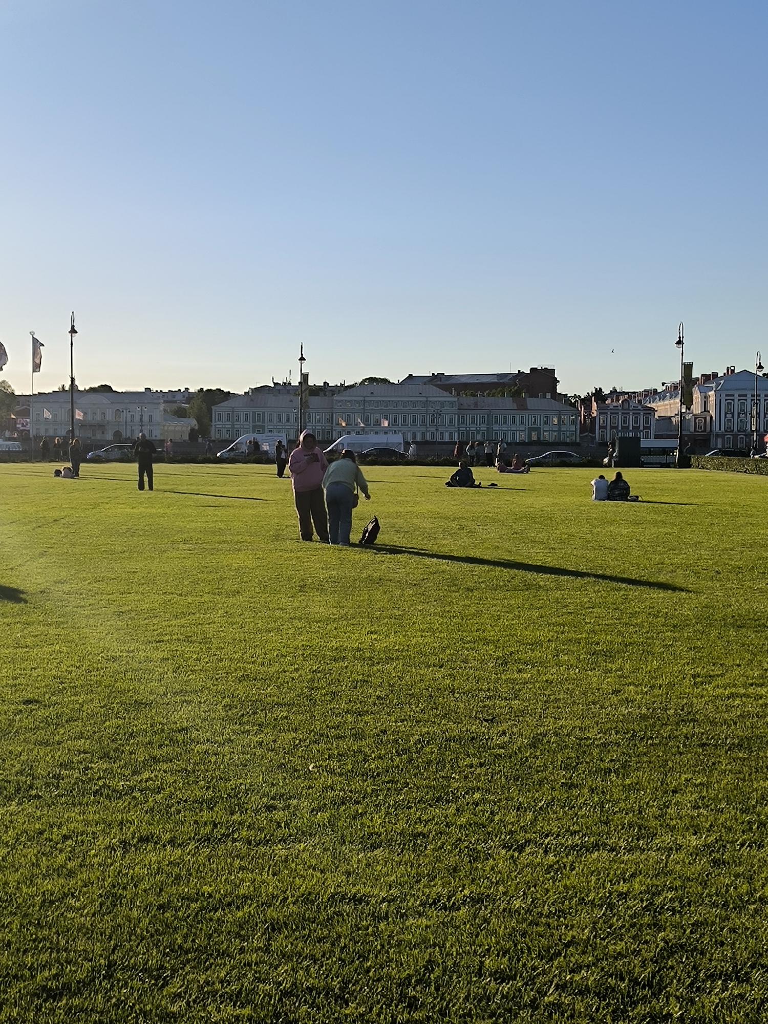 The image depicts a wide expanse of manicured green grass under a clear blue sky, illuminated by the warm glow of late afternoon sun. In the background, the distinctive architecture of Saint Petersburg, Russia, is visible. Buildings with classical facades and mansard roofs line the horizon, with lampposts and flags dotting the scene. Several individuals are scattered across the lawn. In the mid-ground, two women are standing, one in a pink hooded sweatshirt and the other in a light green jacket, with a dark bag between them. Other people are seated or standing further in the background, some appearing to be picnicking. Cars and vans are also present near the buildings, indicating a public urban space. The overall atmosphere is one of peaceful recreation in a historic city setting.