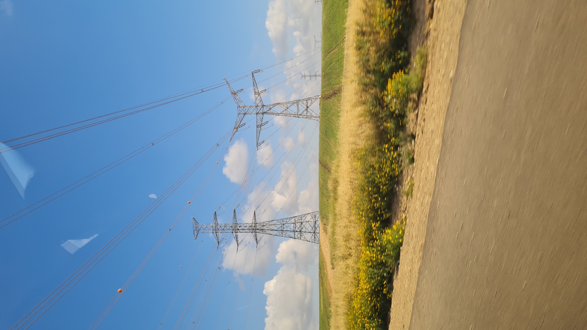 The image shows a view of power lines and a field of yellow flowers next to a road, likely taken from a moving vehicle. The sky is blue with scattered clouds. The power lines run parallel to the road and field. There are orange markers on the lines. The location is likely within or near the Bnei Shimon Regional Council in Israel, given the presence of power lines and agricultural fields which are typical of this area.