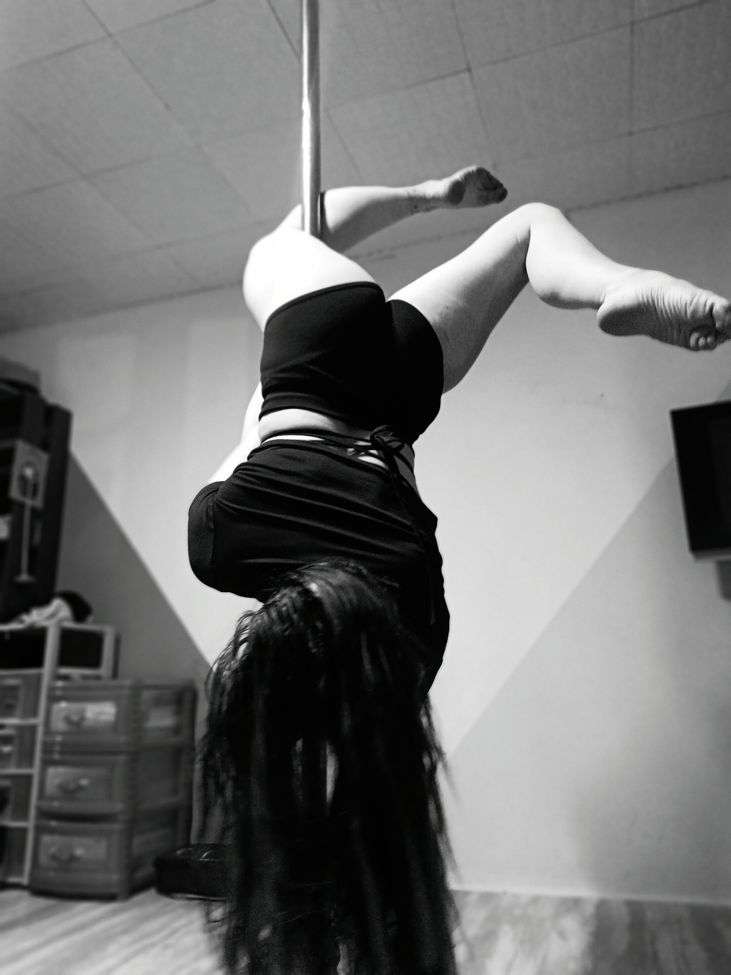 A black and white photograph shows a person suspended upside down on a pole, presumed to be a dance pole, in an indoor setting. The person is wearing dark shorts and a top, with long hair cascading downwards. Their legs are bent and extended upwards, with one arm visible. In the background, there is a light-colored wall, a storage unit with drawers, and a television set. The location appears to be a room in a residence, but the specific city and state are unknown.