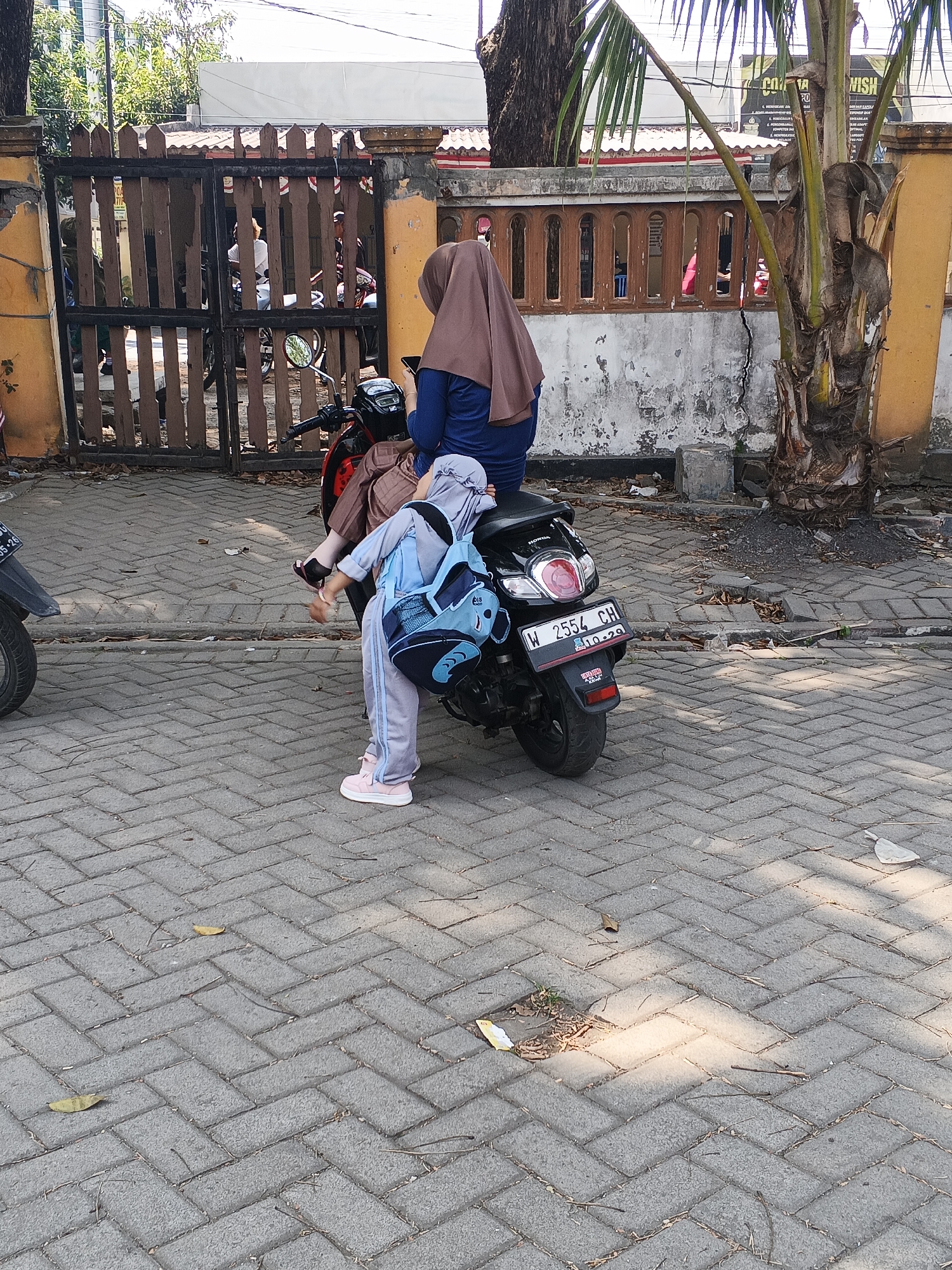In Krian, Indonesia, two individuals are present on a paved area. A woman wearing a brown hijab and a blue top is seated on a black Honda motorcycle. She is holding a mobile phone. A child, dressed in a light blue hijab and a gray tracksuit with pink shoes, is standing beside the motorcycle. The child has a light blue backpack with black trim slung over their shoulders. The motorcycle has a license plate that reads "W 2554 CH". The background features a wooden fence with a yellow-painted pillar and some foliage, including a palm tree. The ground is paved with interlocking bricks, with some fallen leaves and debris scattered on the surface.