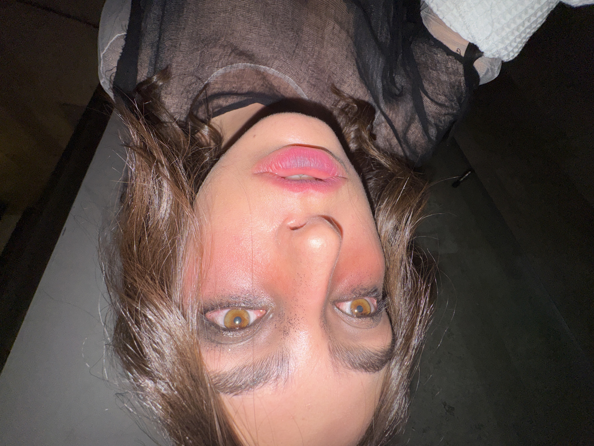 A close-up, upside-down image shows a person with light skin, brown hair, and brown eyes. The person's face appears to be wearing makeup, with dark eyeshadow and pink lipstick. The person is wearing a sheer black top. The background is dark and blurry, obscuring any specific location details. The information available does not include or suggest that the location is Mexico City, Mexico. There are no identifiable actions or interactions within the frame. The image primarily focuses on the person's face and upper torso.