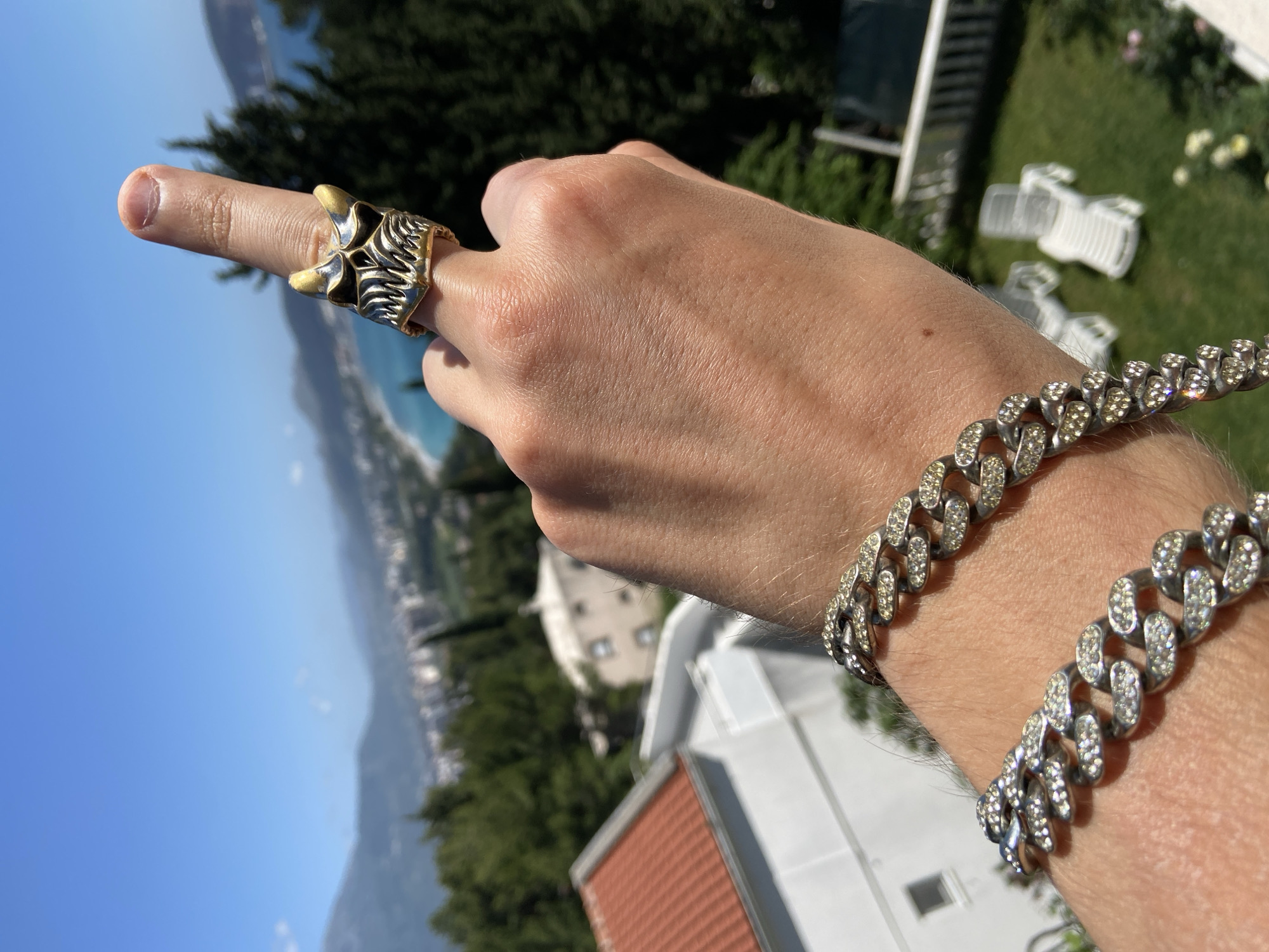 A person's left hand is adorned with a striking silver ring with gold accents, worn on the middle finger. The person is also wearing two silver chain bracelets with small, clear gemstones on their wrist. The person's hand is in front of an outdoor scene, likely in the city of Šušanj, Montenegro.
In the background, there is a view of the Adriatic Sea coastline, mountains, and buildings. The sky is a clear, bright blue. In the foreground, there is a lawn, trees, white lawn chairs, and buildings with red tile roofs, typical of the architecture in that region. The person is most likely standing on a balcony or in a yard overlooking the sea. The image is taken during the daytime under clear and sunny weather conditions.