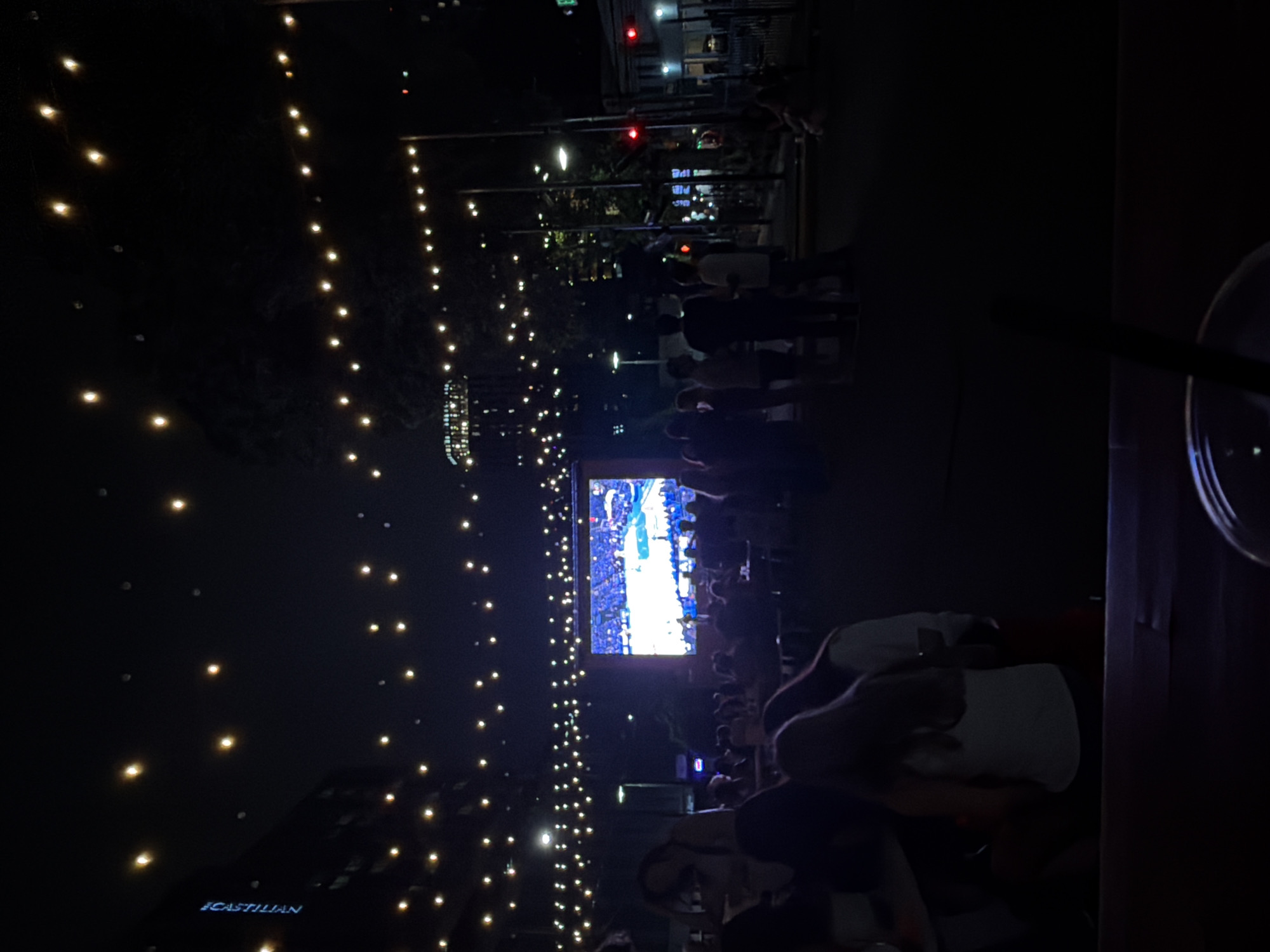 The image shows an outdoor gathering at night in Austin, Texas. A large screen displays a basketball game, surrounded by people watching. String lights hang overhead, illuminating the scene. The building "Castilian" is visible in the lower-left corner. A drink with a straw sits on a table to the right.
