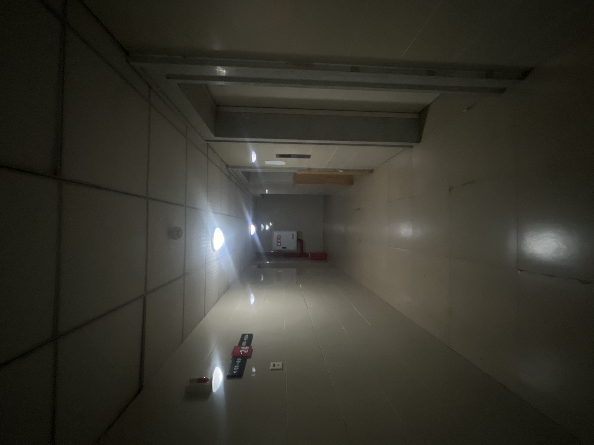 The image depicts a dimly lit, tiled hallway, likely within a residential or commercial building in Jiulongpo District, China. The hallway extends into the distance, with fluorescent lights on the ceiling providing illumination. On the left wall, a sign indicates room numbers "01-09" and "24," along with an emergency call button. Further down the hallway on the right, a fire alarm box is visible on the wall. The floor is made of large, light-colored tiles, reflecting the overhead lighting. The overall impression is one of an unremarkable, functional interior space.