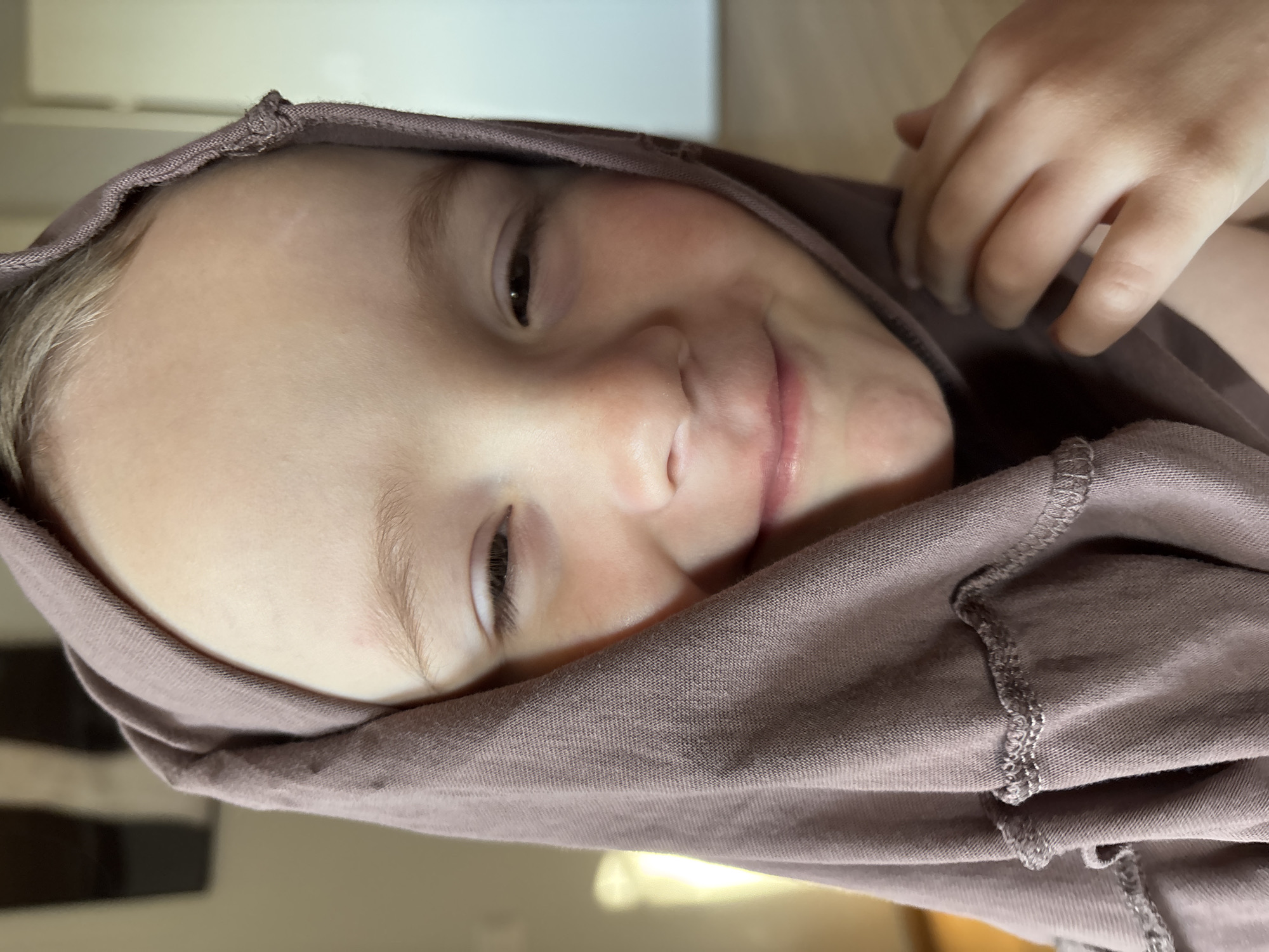 A close-up, slightly rotated, indoor portrait shows a child with fair skin and light brown hair. The child is partially obscured by a mauve-colored cloth that drapes over their head and around their face, with a hand visible above the cloth on the right side of the frame. The child's eyes are dark, and they have a slight smile. The background is out of focus, showing blurred shapes and light colors. The location cannot be determined from the image.