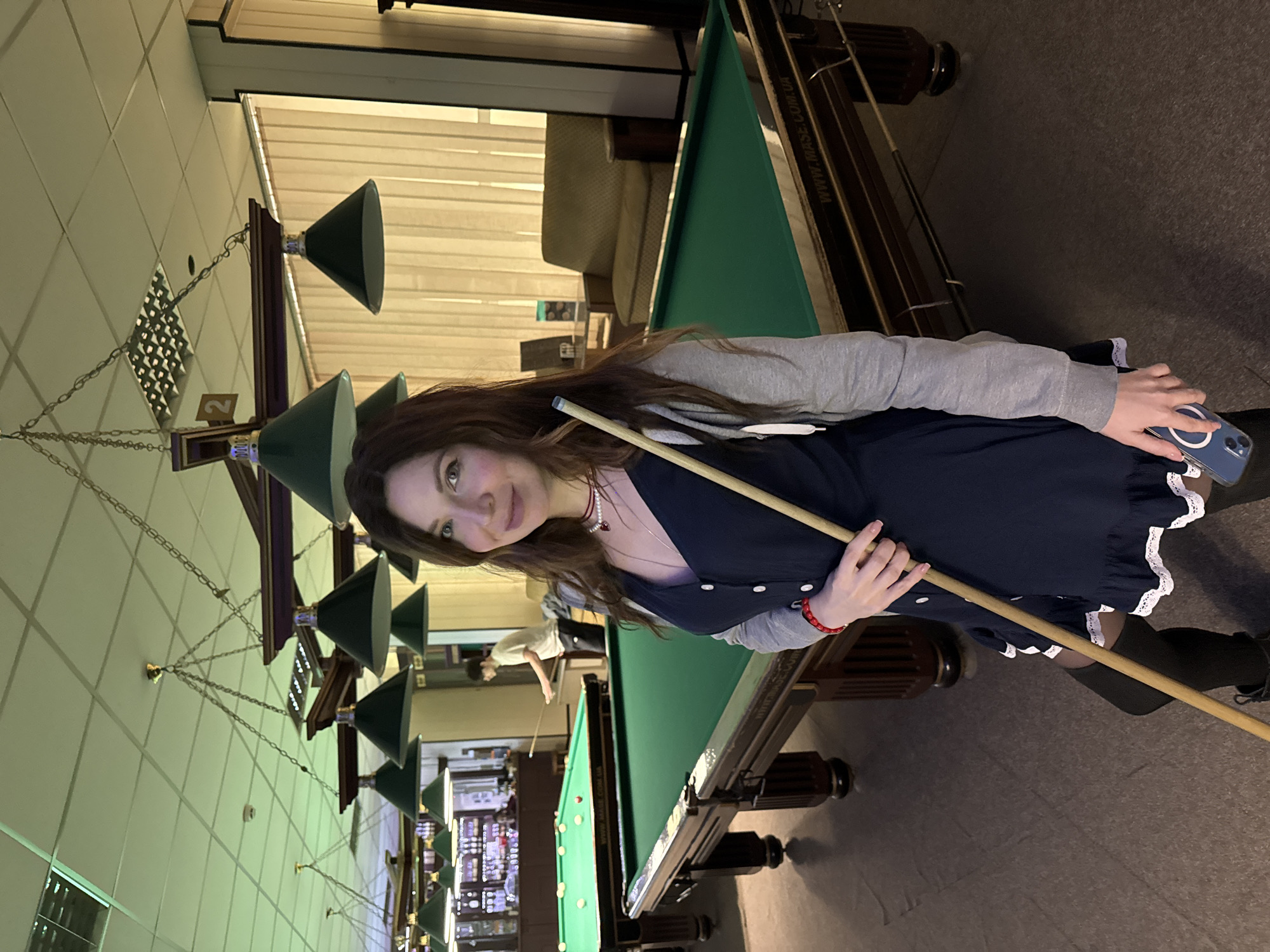 A woman stands next to a billiard table holding a cue stick in what appears to be a billiard hall. The billiard hall is illuminated by a row of conical light fixtures suspended from the ceiling by chains. There are other billiard tables visible in the background, including one where another person is playing. A bar with illuminated shelves can be seen in the far background. The woman wears a dark dress with white trim, a light gray sweater, dark socks, and dark shoes. She is holding a smartphone in her left hand. The billiard table is covered in green felt and has several billiard balls on it. Based on the inscription "WWW.MAZE.COM.UA" on the side of the table, it is possible this scene could be in Ukraine. Kryvyi Rih is a city in Ukraine, so it is possible that the image was taken there.