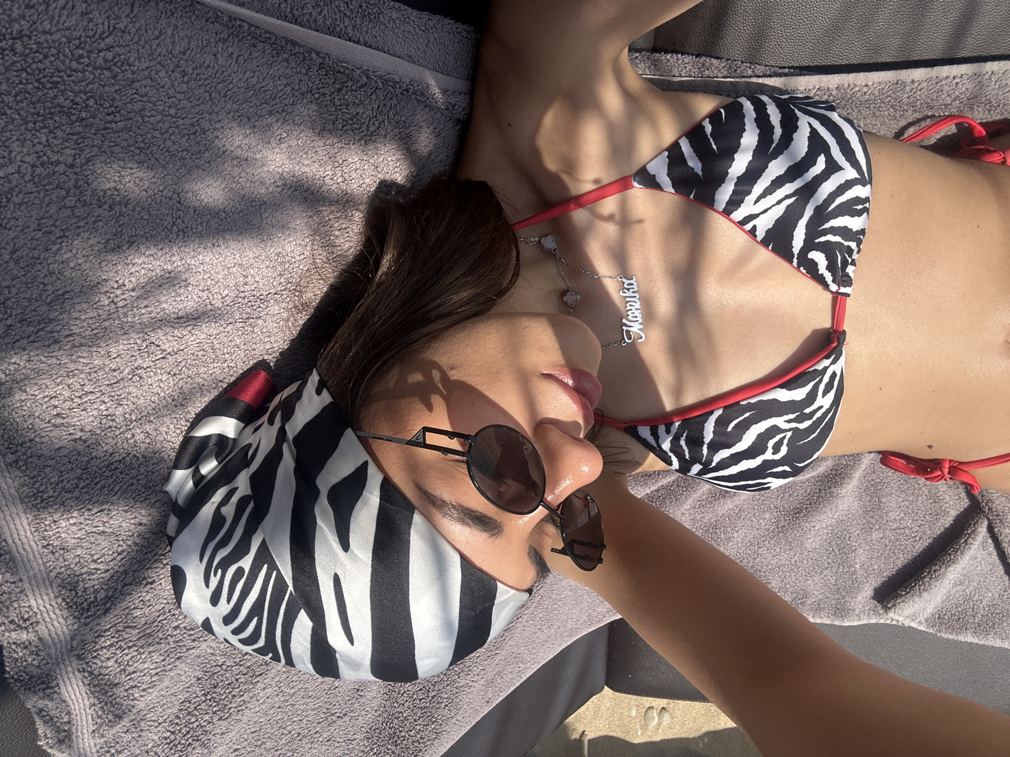 A young woman lies on a towel on a lounger at Ofrynio Beach, Greece, enjoying the sun. She is wearing a zebra-print bikini and a black and white striped headscarf. Her sunglasses have round, dark lenses and thin black frames. A silver necklace with a nameplate that reads "Marlika" and two clover-shaped pendants hangs around her neck. Sunlight casts dappled shadows across her body and the towel.