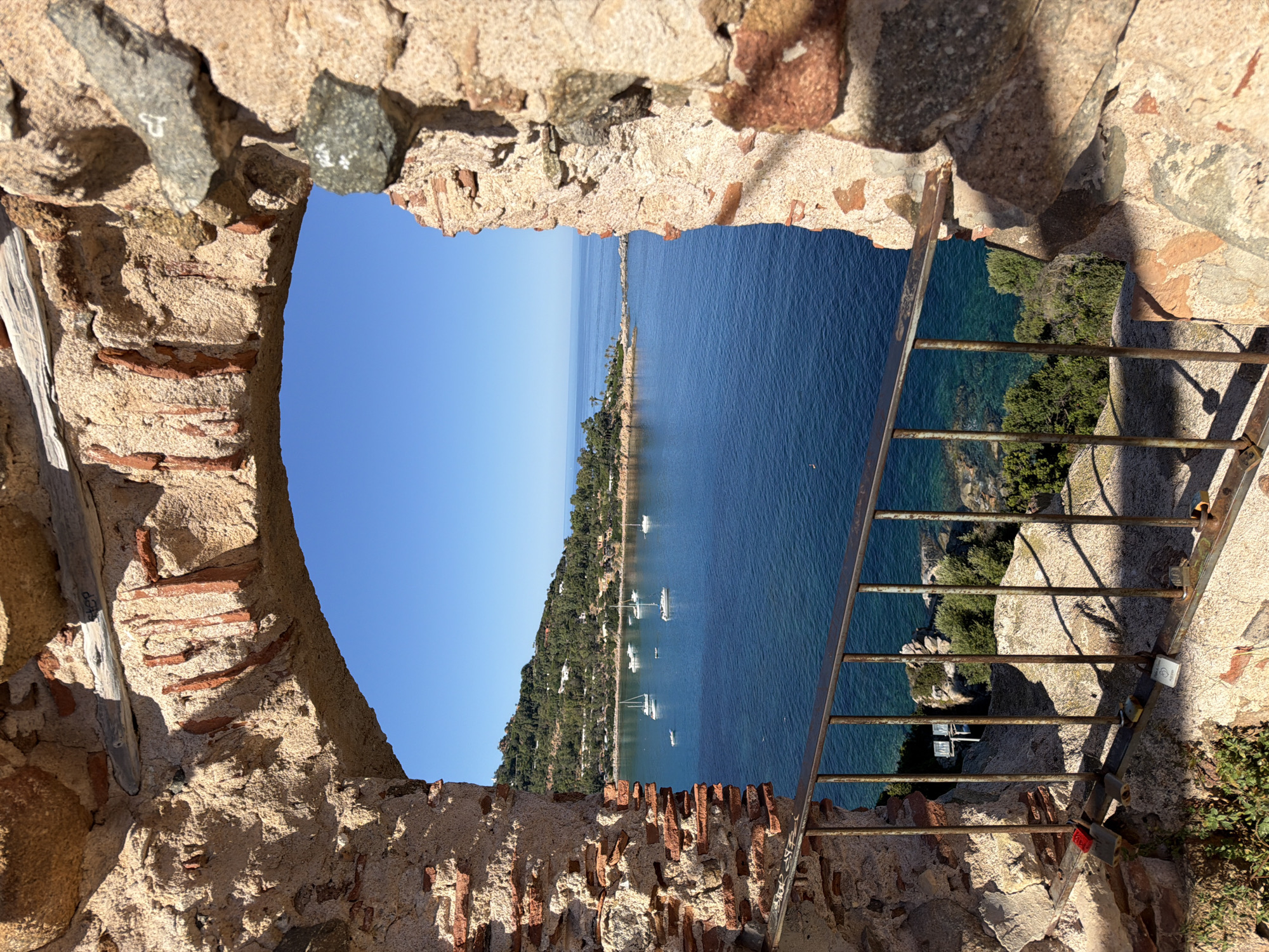 The image shows a view through an arched stone opening overlooking a blue sea with several sailboats. The stone structure is rough and weathered, made of large rocks and smaller red bricks. A metal railing runs along the bottom of the opening. In the distance, a green, hilly coastline dotted with white buildings can be seen. The sky is clear and blue. The scene appears to be in the vicinity of Tortolì, Italy, likely a viewpoint from a historic structure or fortification.