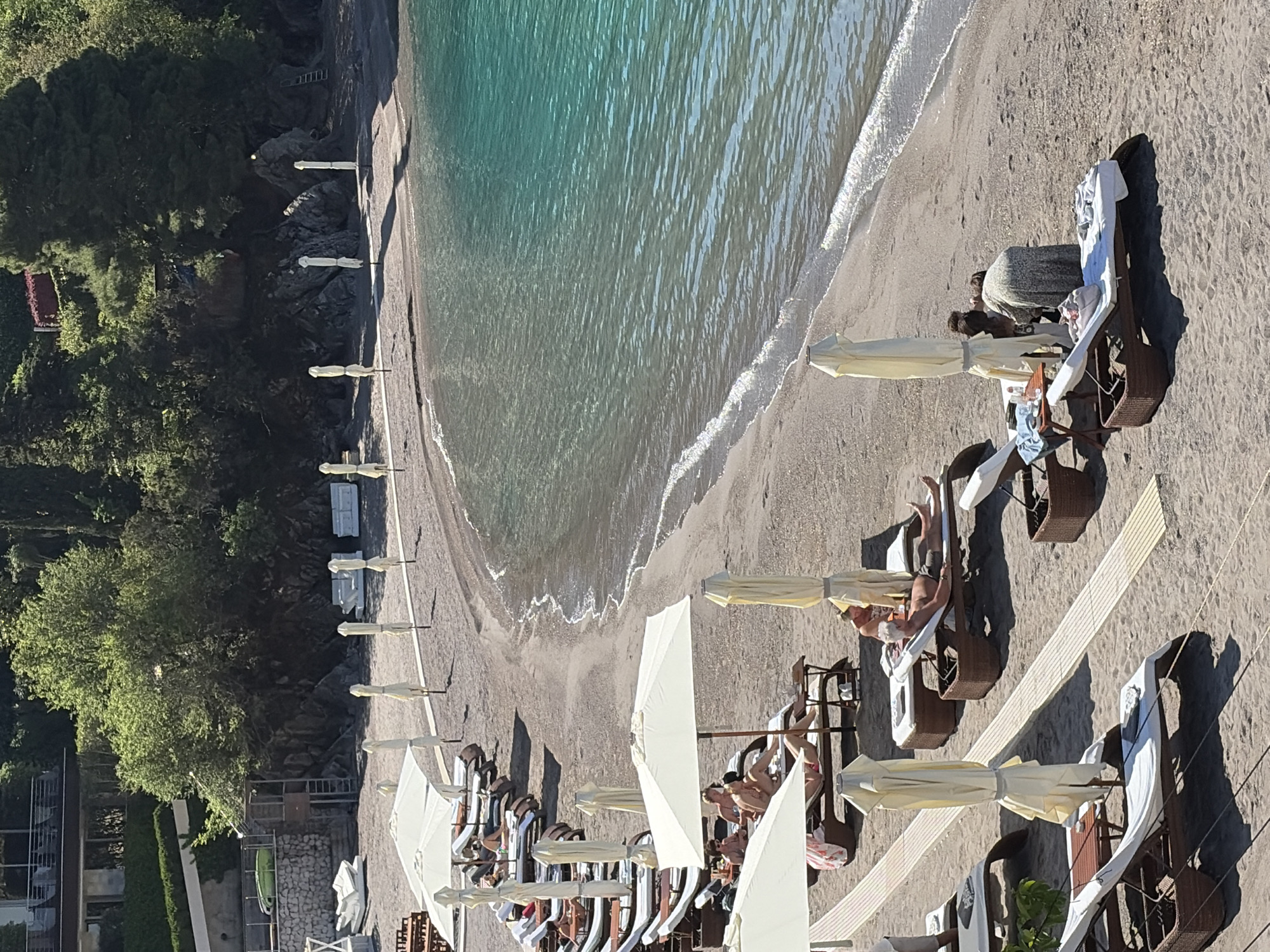 The image shows a beach in Budva, Montenegro. The beach has dark sand and turquoise water. A row of beach chairs and umbrellas are lined up along the beach, with people lounging in some of them. On the left side, a cliff covered in greenery borders the beach. The image is oriented vertically and appears to have been taken from an elevated position.