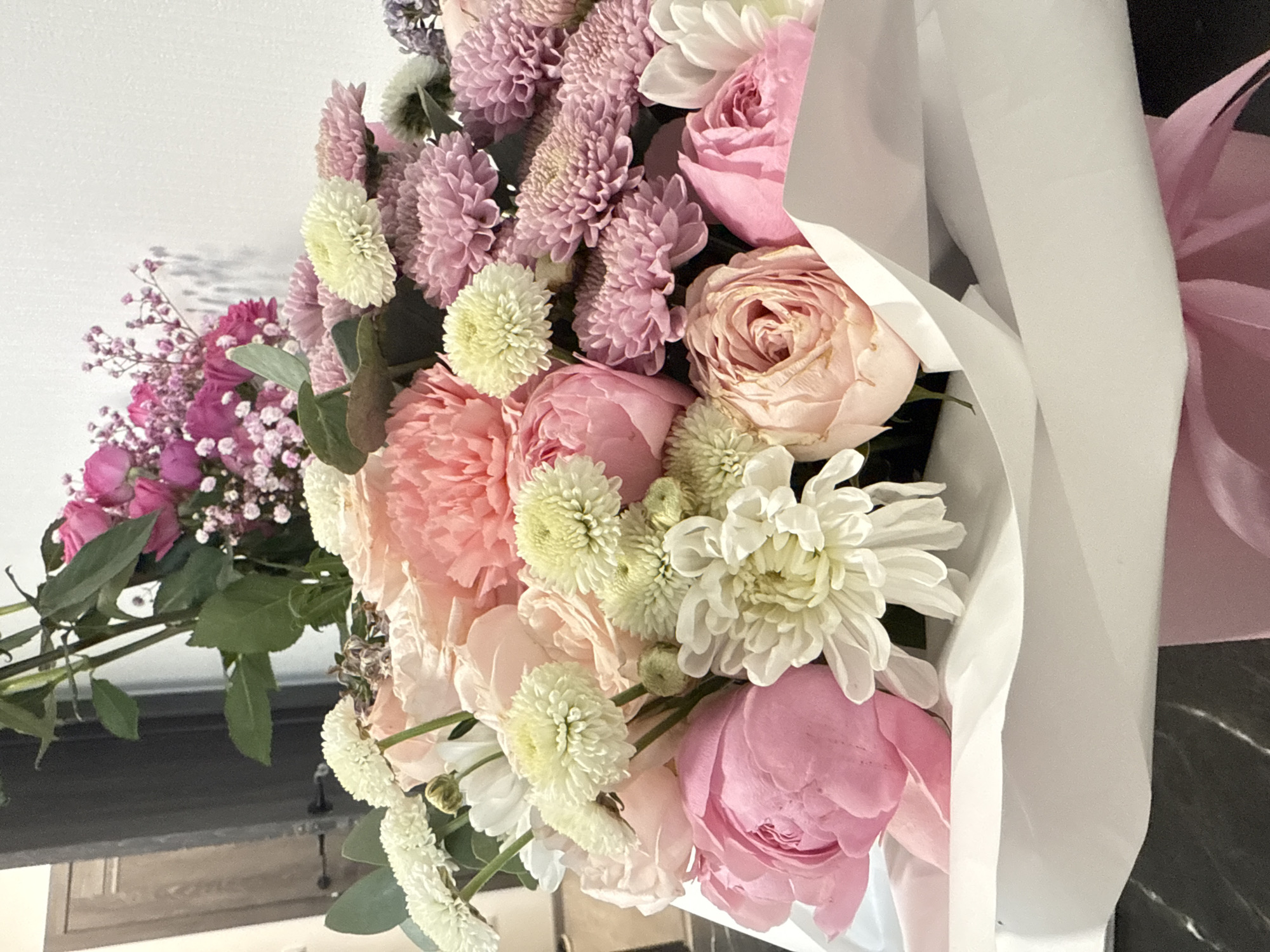 The image is a close-up of a bouquet of flowers. The bouquet includes various types of flowers in shades of pink, white, and light purple. Some of the flowers appear to be roses, peonies, and chrysanthemums. The bouquet is wrapped in white paper with a pink bow. The location of the image is not specified, and there is no information suggesting it is in Saint Petersburg, Russia. The image does not include any people, actions, or interactions. The environment consists of a partially visible countertop and wall in the background.
