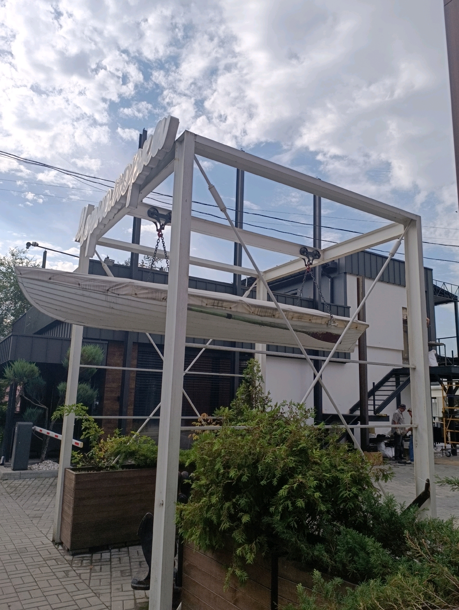 This image depicts the exterior of a building with a unique architectural feature: a white boat suspended from a metal frame above a paved area. The boat is positioned horizontally and appears to be a decorative element. The building itself has a modern design with dark siding and white accents, and features an outdoor staircase. Lush green bushes and planters are visible in the foreground, adding a touch of nature to the urban setting. The sky above is partly cloudy. A barrier arm for vehicle control is also present, suggesting this is an entrance to a property. The scene is captured on a cloudy day, and the overall impression is one of contemporary design blended with unusual artistic elements. It's possible this is a restaurant or a business with a distinct aesthetic.