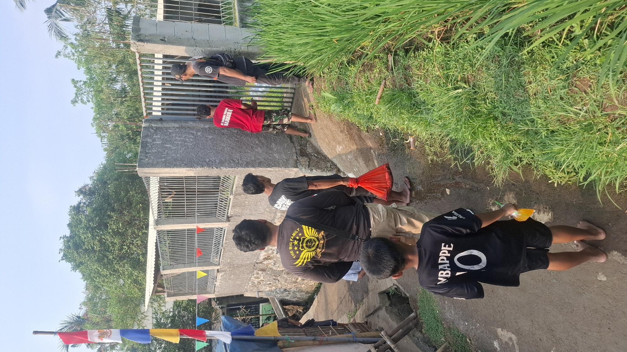In the city of Rahong, Indonesia, a group of men and a boy are gathered in an outdoor alleyway. The ground is damp and uneven, with patches of grass and vegetation lining the right side of the image. To the left, a steep embankment is visible, with a weathered stone wall and a metal fence. Colorful pennant flags are strung across the upper left portion of the scene, and several Indonesian flags are affixed to a pole.

In the foreground, a young boy, barefoot, wears a black t-shirt with "MBAPPE" and the number "10" emblazoned on the back. He holds a small orange object in his right hand. Behind him, a man in a black long-sleeved shirt with yellow winged graphics is visible from the back. Further back, another man in a black t-shirt is seen from the side, carrying a bright red plastic bag.

To the right, a man in a red t-shirt with the text "BANDINGKAN NARKOBA" stands in front of a metal gate. Next to him, another man wearing a black t-shirt and a black cap is also facing the gate. The gate is part of a concrete structure that appears to be a building or enclosure. The sky above is clear and bright blue, suggesting a sunny day. The overall impression is of a community gathering or event taking place in a semi-rural or densely populated urban environment.