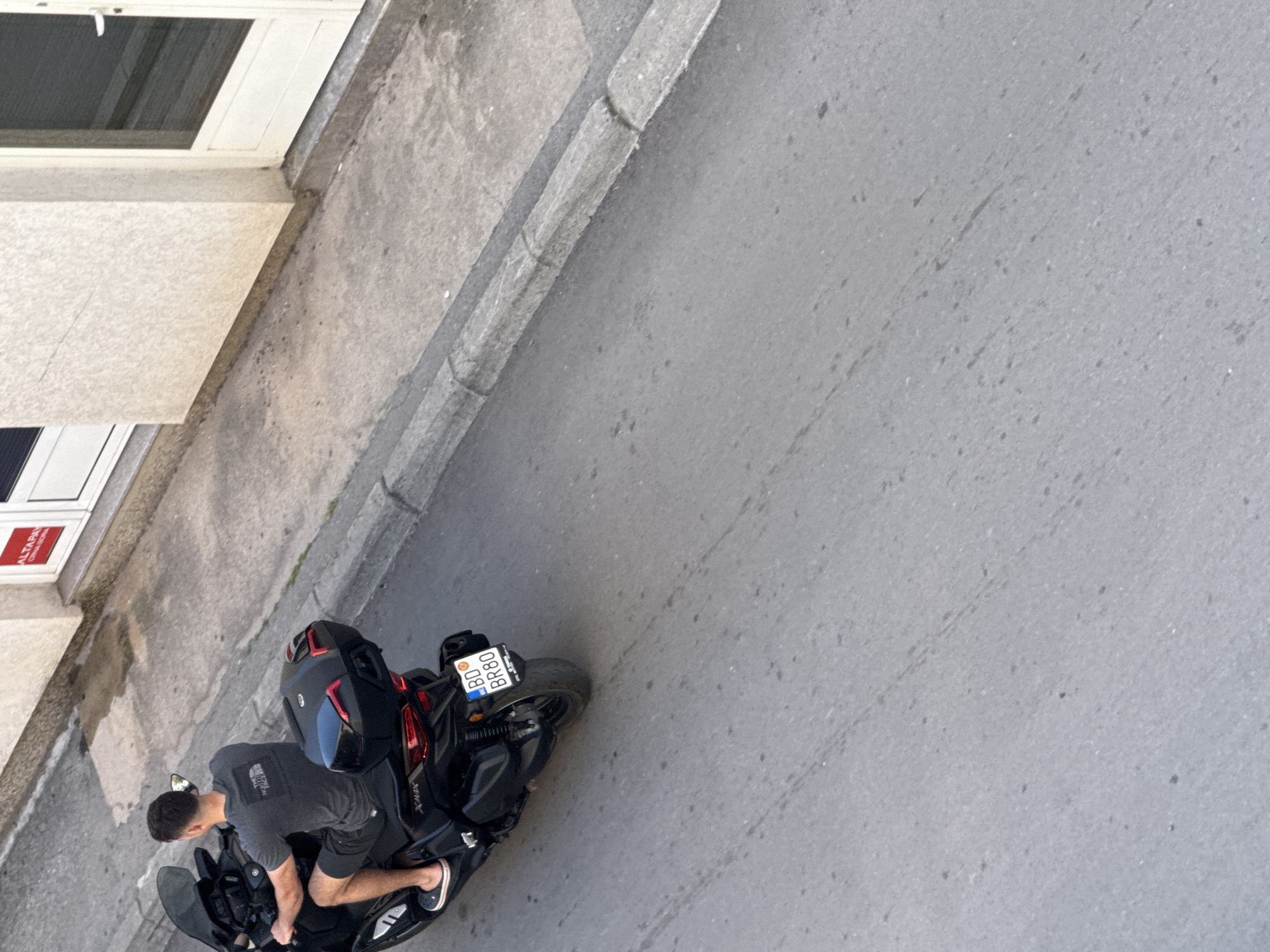 The image depicts a man on a black Yamaha XMAX motorcycle parked along a street. The license plate reads "BD 80 BR80". The man is wearing a grey shirt, black shorts, and black shoes. The motorcycle has a black helmet and storage compartments. The surrounding environment includes a building with windows and a street. The location is likely a street in Budva, Montenegro, based on the license plate which indicates the city of Budva.