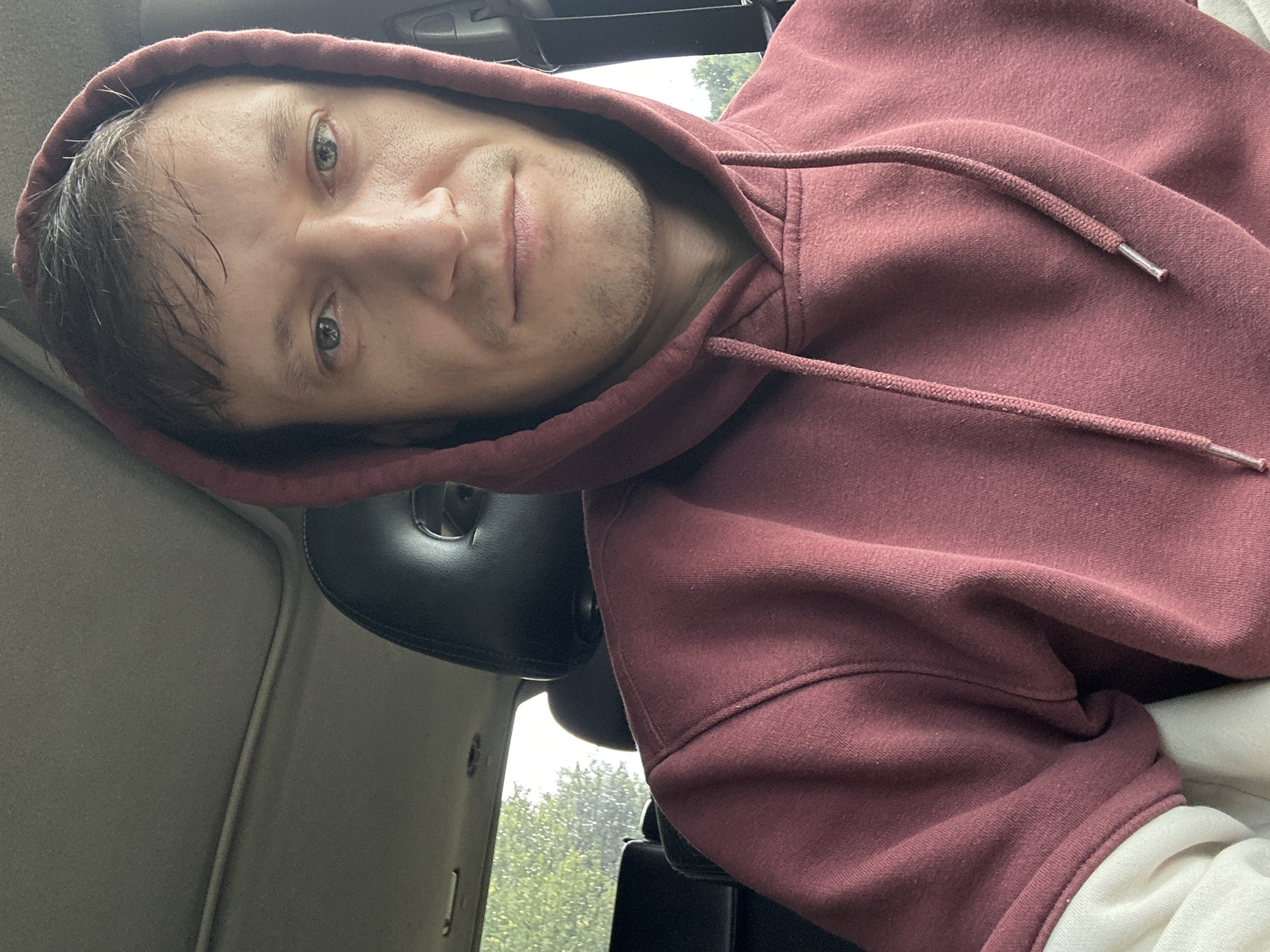 A man with short, dark hair and light-colored eyes is pictured from the chest up, looking directly at the camera. He is wearing a dark red hooded sweatshirt with a cream-colored undershirt visible at the hem. The background shows the interior of a vehicle, including a black headrest, a sunroof, and a window with trees and foliage visible outside. The man appears to be in the driver's seat of the vehicle, suggesting he is located in Makiv, Ukraine, as indicated by the provided context. The overall lighting suggests it is daytime.