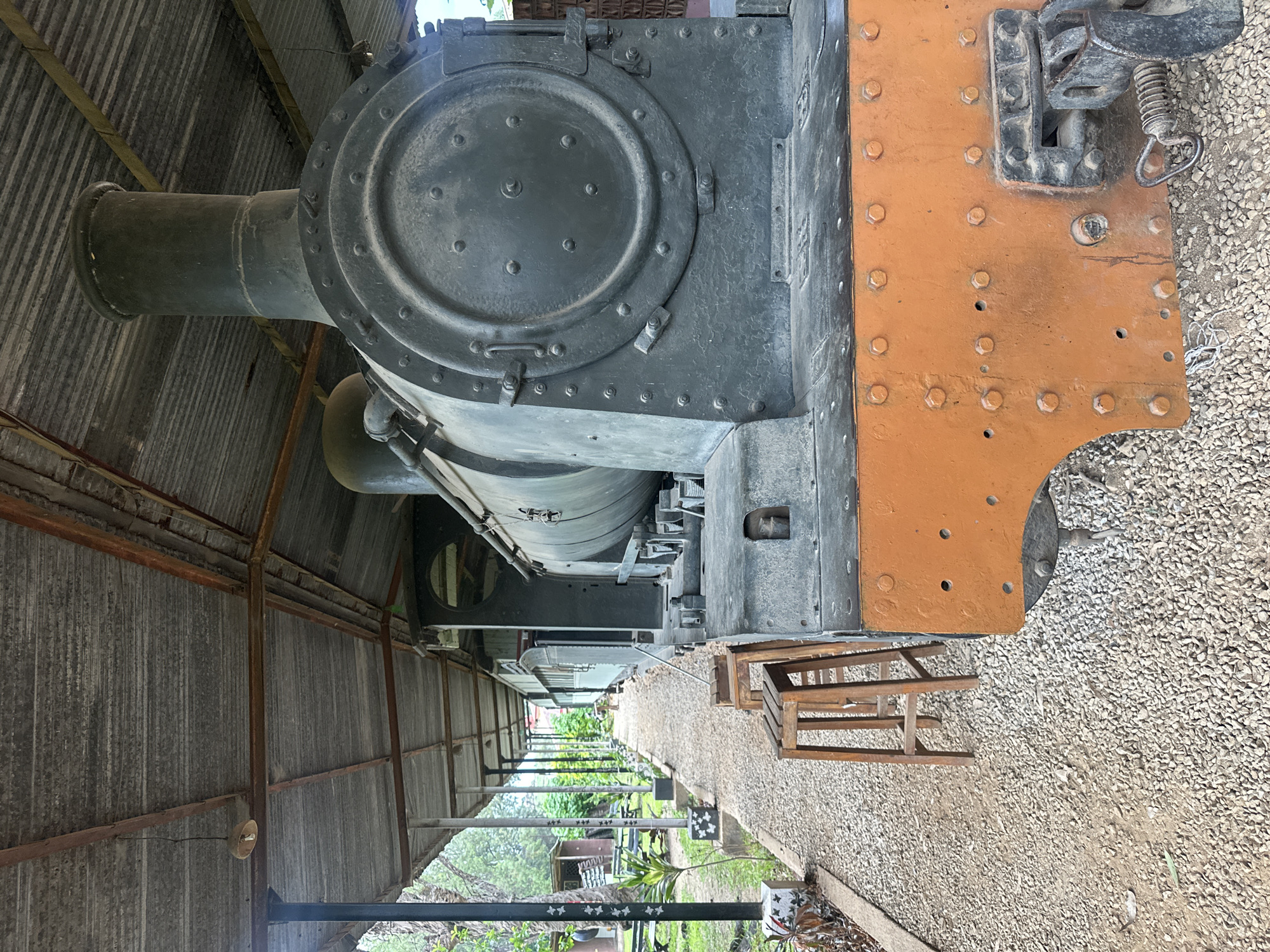 This image captures a vintage steam locomotive housed within a corrugated metal shed, likely at a historical site or museum in Jos, Nigeria. The photograph is taken from the front-right side of the locomotive, looking along its length.
The primary subject is a dark-colored steam locomotive, specifically its front section. A prominent, large, circular access panel on the front of the boiler is secured with numerous rivets. Above this, a wide smokestack extends upwards. The front buffer beam of the locomotive is painted a distinct orange/rust color, also held together by many bolts and rivets, and features a robust coupling mechanism with a spring. The locomotive's metallic body shows signs of age and wear.
The setting is an open-sided shed with a roof and walls constructed from corrugated iron sheets, supported by metal beams. The shed provides shelter for the locomotive. The ground beneath the locomotive and extending into the background is covered with light-colored gravel, characteristic of a railway track bed or platform. In front of the locomotive, on the gravel, there's a simple wooden stool or crate.
Beyond the shed structure and the gravel, the background reveals lush green foliage, indicating an outdoor environment with trees and plants. The lighting suggests it is daytime, with natural light illuminating the scene. No people are visible, and the locomotive is stationary, implying it is either on display or in storage. There is no visible text in the image. The scene evokes a sense of industrial history and preservation.