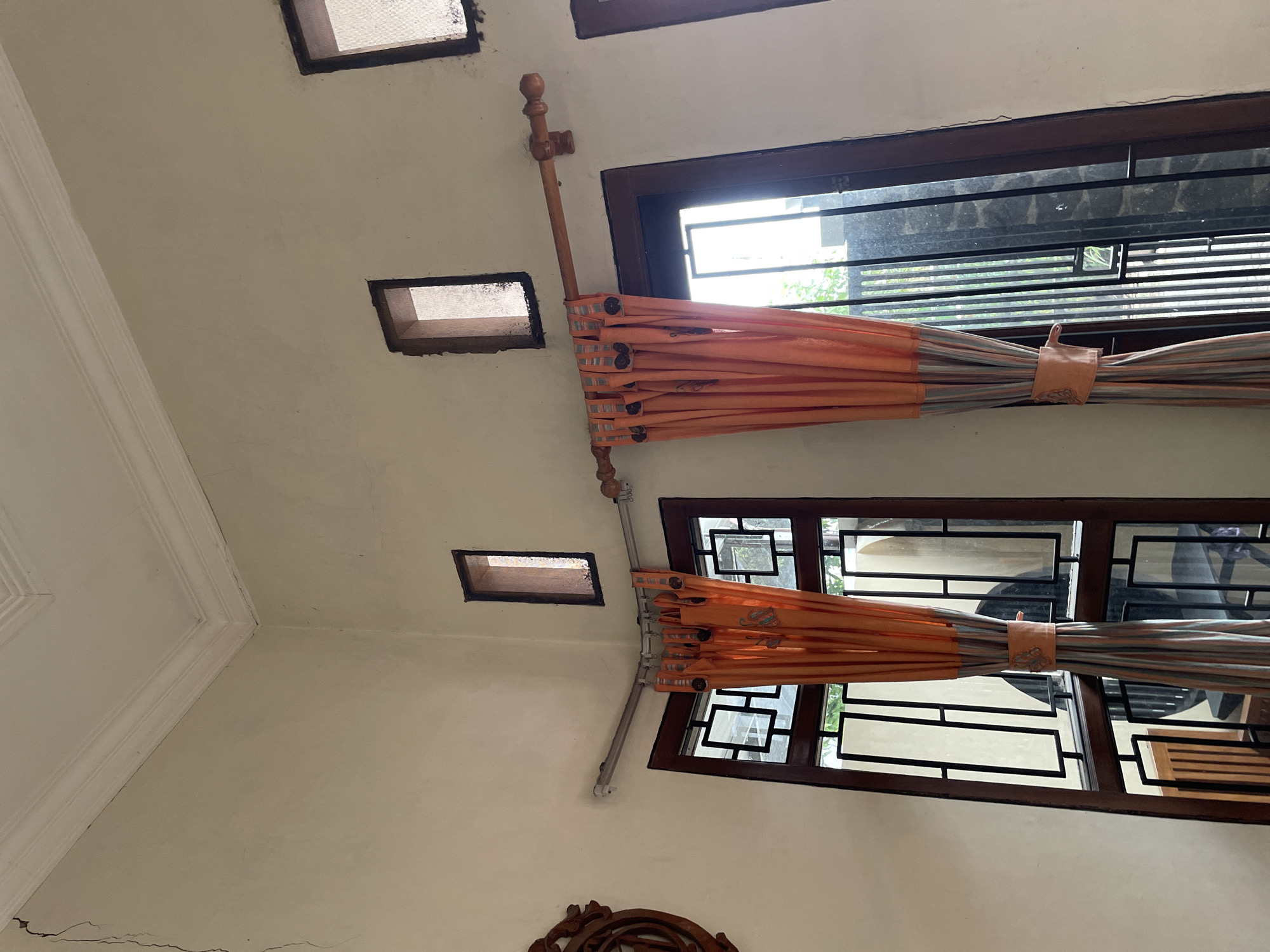 The image depicts an interior corner of a room, likely a residential dwelling in Pekalongan, Indonesia. The walls are painted a light cream color and show signs of wear and cracking, particularly a prominent vertical crack extending from the ceiling down the left wall. The ceiling features decorative molding.
In the corner, two windows are visible. The left window has a wooden frame with a grid-like pattern of dark metal bars. An orange curtain with a striped pattern is drawn across this window. The right window is a larger, dark-framed window or door, also with metal bars. A similar orange curtain is drawn across this opening. Both curtains are suspended from wooden curtain rods.
Above the windows, there are two small, rectangular openings in the wall, characteristic of ventilation or decorative features often found in Indonesian architecture. A decorative wooden carving is partially visible on the far left of the image. The overall impression is of a somewhat aged interior, possibly in need of renovation.