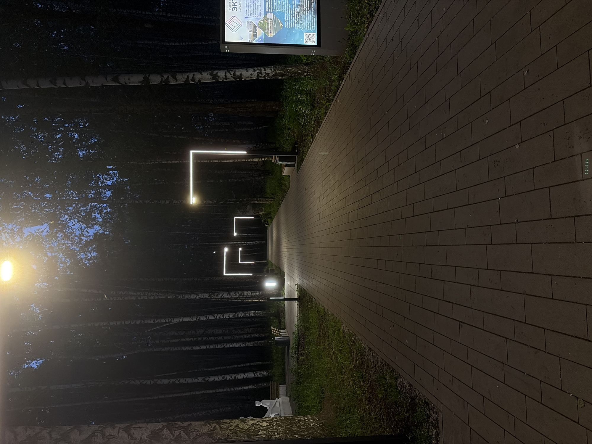 The image captures a well-lit, paved pedestrian pathway at night, receding into the distance. The pathway is made of rectangular, light-colored paving stones and stretches from the foreground towards the upper-center of the frame.
To the left of the path, a dense line of trees, predominantly birches with their characteristic white bark, forms a dark backdrop. A bright, warm-toned street lamp illuminates the immediate foreground on the left, casting a glow onto the trees. Further along the path, several modern, L-shaped or rectangular outline lights emit a cool white luminescence, accentuating the path's linearity. A dark wooden bench is visible under one of these modern lights. In the foreground on the left, a light-colored, possibly white, abstract sculpture or figure is situated among the trees.
To the right of the path, a strip of grass separates the walkway from what appears to be part of the park's landscape. In the foreground on the right, an illuminated information stand or digital display board is visible. The stand features Cyrillic text, with prominent words at the top reading "ЭКО-ПАРК" (Eco-Park) and "УРАЛО...". It also displays images of modern architecture, a QR code, and smaller descriptive text. Further down the path on the right edge, a small panel with several green indicator lights is visible.
The scene depicts a peaceful and modern urban park or pedestrian zone, designed for evening strolls. The clear night sky is suggested by the crispness of the artificial lighting. No individuals or specific activities are visible, contributing to the serene atmosphere.