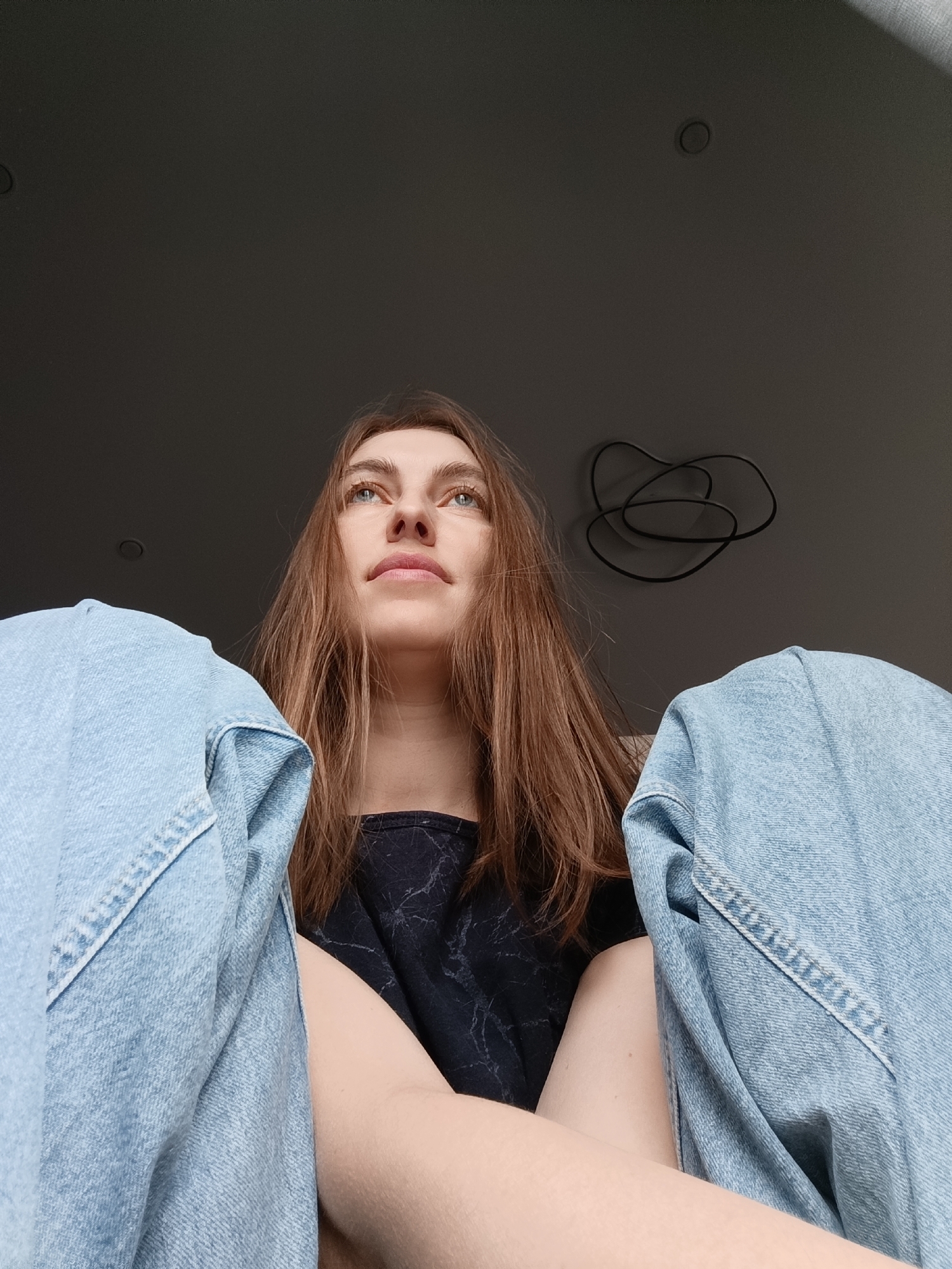 The image depicts a woman with long brown hair and blue eyes looking upwards. She is wearing a dark top and light blue denim jeans. The perspective is from a low angle, showing her from the chest up with her legs positioned on either side of the frame. The background is a grey ceiling with a tangled black cable fixture. The location is presumed to be Chaikovsky, Russia. No actions or interactions are visible; the subject is simply looking up.