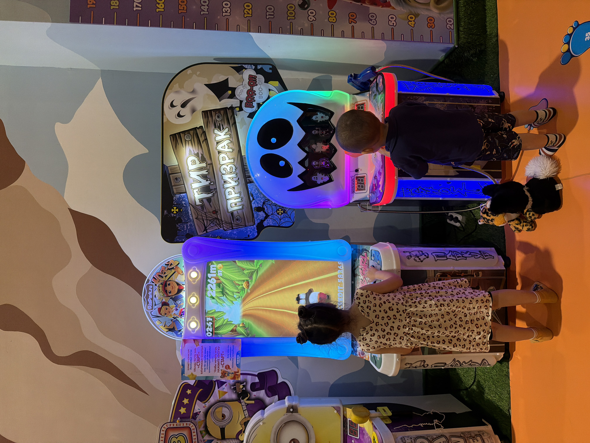 In an indoor amusement setting, two children, a girl and a boy, are engrossed in playing arcade games. The girl, facing away from the camera, wears a leopard-print dress and plays a game featuring animated characters on a bright screen. The boy, also facing away, is dressed in a dark t-shirt and patterned shorts, playing a game with a ghost theme, illuminated by colorful LED lights. A stuffed toy animal is positioned on the floor between the two arcade machines. The background wall features a decorative mural with a wavy pattern and a height chart indicating measurements in centimeters. The floor is covered with orange and green carpeting. The overall scene depicts a typical children's entertainment area.