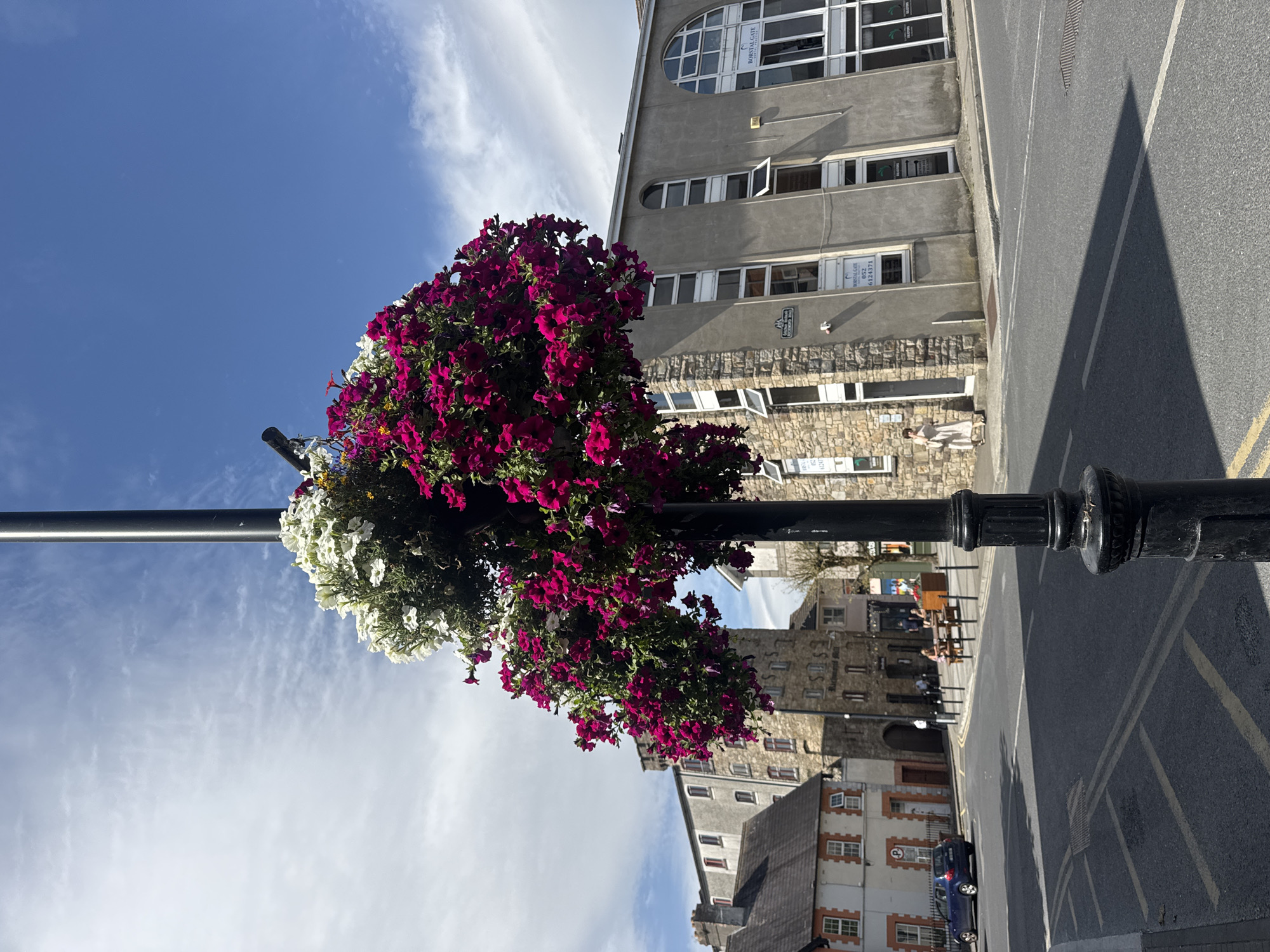 The image depicts a street scene in Clonmel, Ireland. In the foreground, a black lamppost adorned with a large, overflowing hanging basket of vibrant magenta petunias and white flowers stands prominently. The lamppost has a decorative base. Beyond the lamppost, a paved street with yellow double lines is visible, with a shadow cast from the lamppost and flowers stretching across it.
To the left, a blue car is parked on the side of the street. Behind the car is a building with a distinctive orange-brick facade and white-framed windows. Further back, a multi-story stone building with arched windows dominates the middle ground. A woman in a light-colored dress is walking on the sidewalk in front of a modern, grey building on the right side of the street. This grey building has large windows and signage that reads "BORSTAL GATE."
In the distance, several people are seated at picnic tables outside what appears to be a business or cafe. The sky above is a bright blue with scattered white clouds. The overall impression is a lively urban setting on a sunny day, with a focus on the floral display.