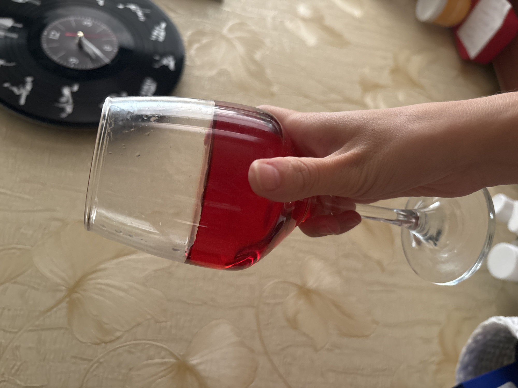 A person's hand holds a wine glass filled with red liquid. The background features a textured wallpaper and a black vinyl record clock with silhouetted figures. The scene is likely indoors, possibly in an apartment in Naberezhnye Chelny, Russia, given the visual cues which suggest a domestic setting.