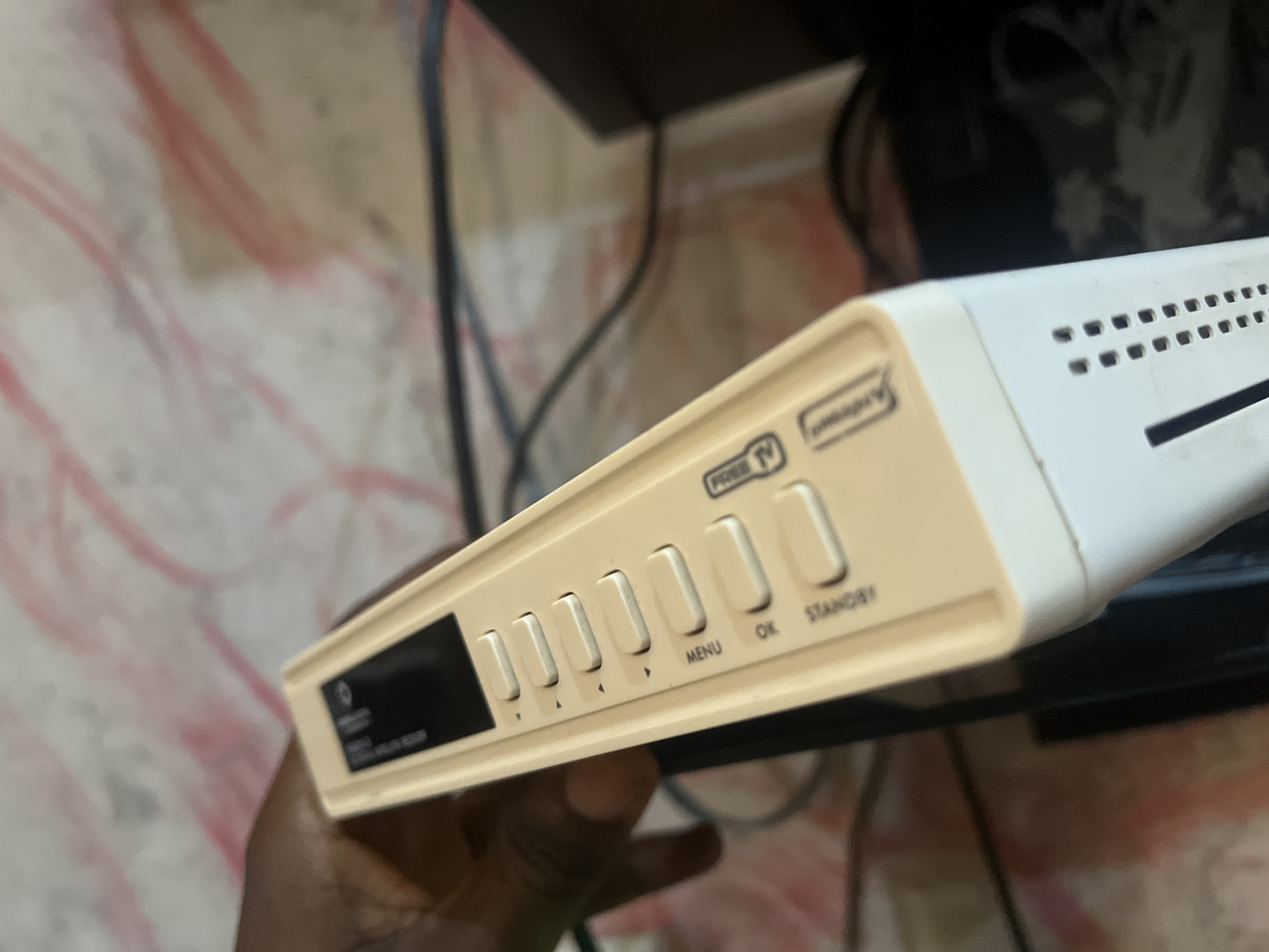 A close-up, angled, eye-level shot shows a person's hand holding a cream-colored satellite receiver box in what appears to be a home environment in Jalingo, Nigeria. The receiver box has a black display screen on the left, followed by several white buttons with icons and labels. The visible labels include "FREE TV," "MENU," "OK," and "STANDBY." The background is blurry, showing a patterned surface that could be wallpaper, and the edges of what seem to be electronic devices and cables. The lighting is moderate, highlighting the textures and details of the receiver box.