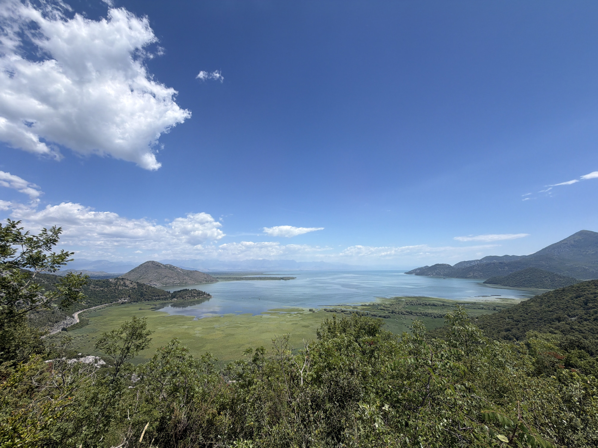 The media file captures a wide-angle view of a serene lake and its surrounding landscape on a clear, sunny day. The scene is dominated by a vast expanse of shimmering blue water, dotted with patches of vibrant green marshland. In the foreground, lush green foliage and trees create a natural frame, obscuring parts of the view and adding depth to the image.
Beyond the lake, rolling hills covered in dense vegetation rise up, their slopes softened by a light haze that suggests distance. The contours of the hills are repeated in the far background, where a range of mountains forms a hazy silhouette against the bright blue sky. A few scattered white clouds drift across the sky, their shapes irregular and wispy, indicating a pleasant, fair-weather day. The sunlight appears to be high in the sky, suggesting it is midday or early afternoon.
The overall impression is one of tranquility and natural beauty. There are no people or discernible objects in the scene, emphasizing the unspoiled, natural state of the location. The absence of man-made structures contributes to a feeling of peaceful isolation. The gentle curves of the shoreline, the varied textures of the vegetation, and the expansive sky create a harmonious and inviting panorama. The location appears to be Kruševica, Montenegro, known for its stunning natural scenery and bodies of water.