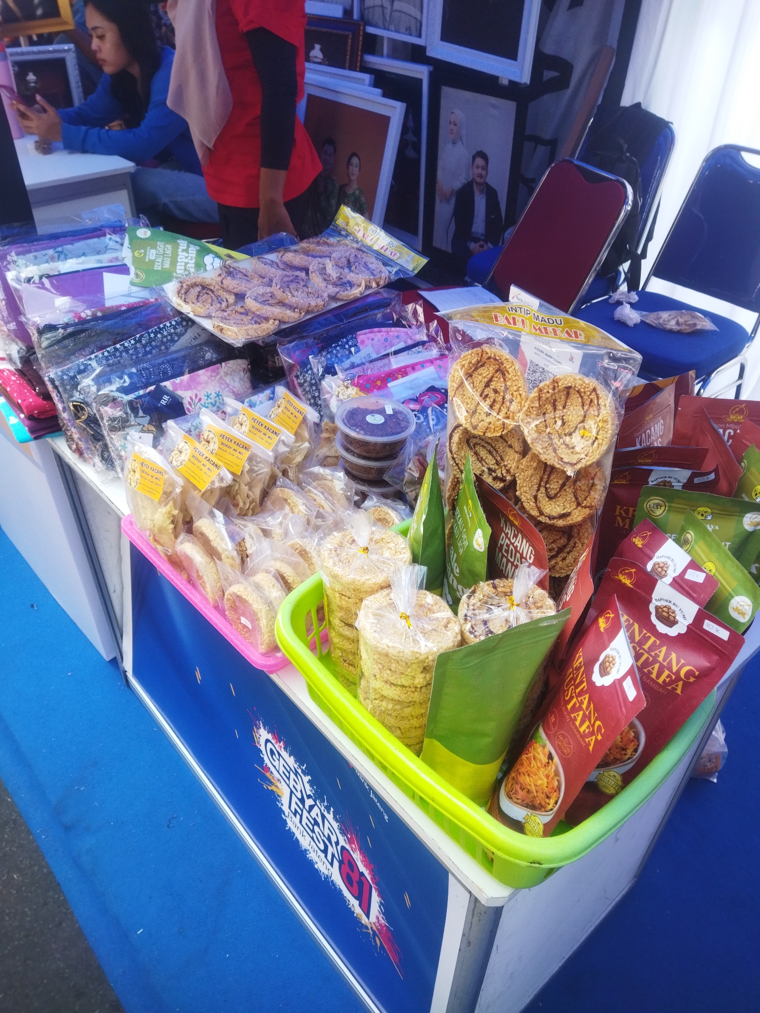 The image shows a street-level view of a stall at the Giripurwo Festival, Indonesia. The stall displays a variety of packaged snacks and food items. In the background, several framed pictures are visible. A woman wearing a blue jacket is seated at a table to the left, looking at her phone. Another person in a red shirt and a headscarf stands behind the table, attending to the stall. The stall itself is adorned with a banner that reads "GEBYAR FEST 81 BANK JATENG." The foreground features several plastic baskets filled with packaged snacks, including what appear to be crackers and other crispy food items. The overall scene depicts a bustling festival environment with commercial activity.