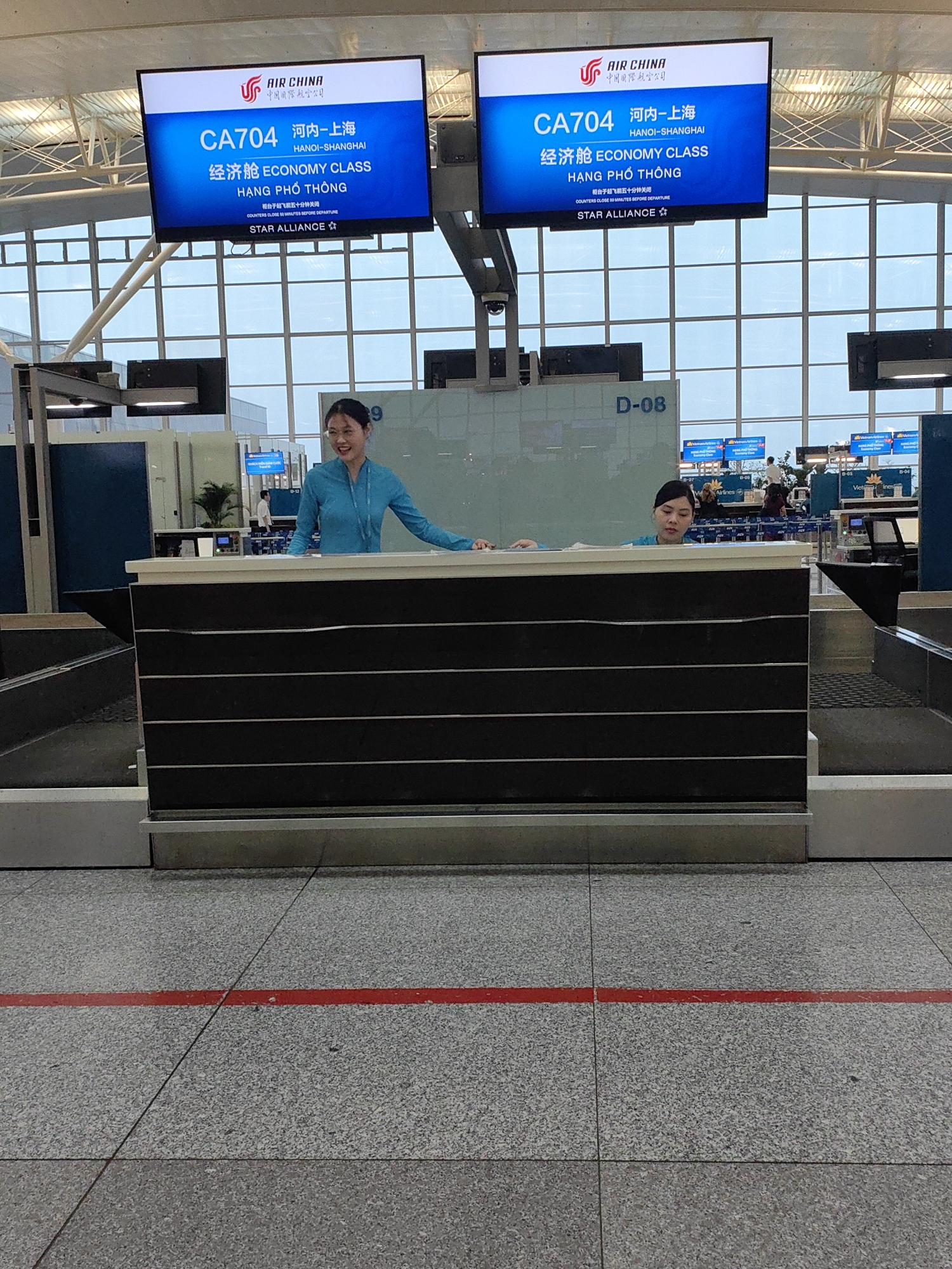 The image shows two women wearing blue uniforms standing behind a check-in counter at an airport. Above them are two digital displays showing flight information for Air China flight CA704 from Hanoi to Shanghai. The displays indicate the flight is for economy class passengers. The counter is labeled "D-08". A red line is painted on the floor in front of the counter. The scene appears to be in an airport terminal. The context from the displays shows this is in Hà Nội, Vietnam.