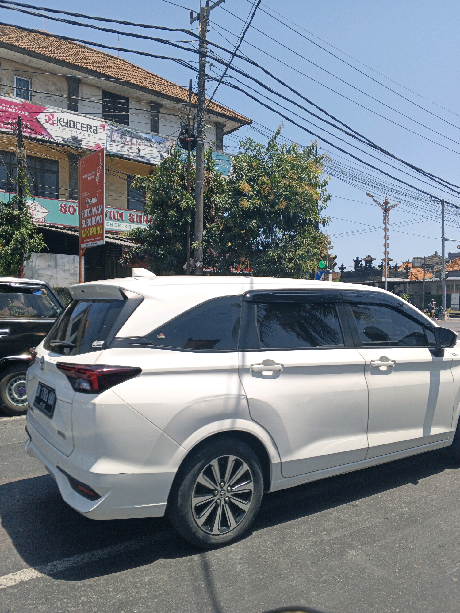 The image depicts a street scene in Denpasar, Indonesia, under a clear blue sky. A white SUV is prominently featured in the foreground, traveling on a paved road. The vehicle has tinted windows and a spoiler on the rear. To the left of the SUV, a black vehicle is partially visible. In the background, a multi-story building with a tiled roof stands, displaying prominent signage for "KYOCERA DOCUMENT SOLUTIONS". Below this, a red vertical sign advertises "Spesial SOTO AYAM SUROBOYO CAK IPUNG". A tree with yellow flowers is visible to the left of the building, and further in the background, traditional Balinese architecture with tiered roofs can be seen. Overhead, a complex network of electrical wires crisscrosses the sky, and a traffic light shows a green signal. The overall environment suggests a sunny day in a bustling urban area.