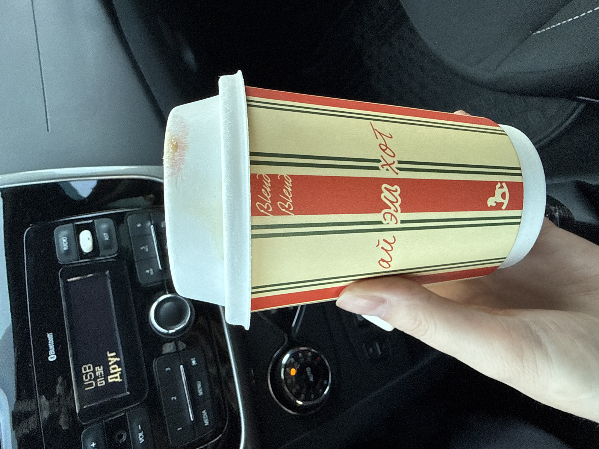 A tan and red striped disposable coffee cup with a white lid rests in a person's hand inside of a car. There is a light pink mark on the lid. The car's radio panel displays "USB 01:32 Друг" and the word "Bluetooth". The cup's text includes "Blend" and "ай эм хот". The cup also has a logo of a rocking horse. The location of Izhevsk, Russia cannot be confirmed by the image.