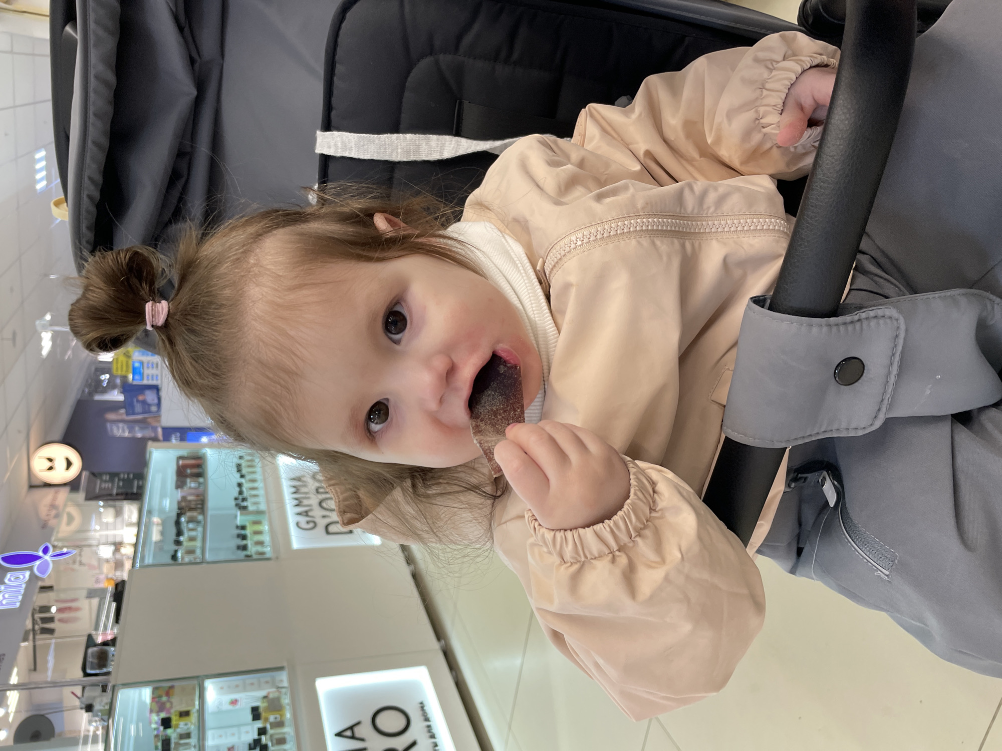 An infant or toddler with light brown hair tied in a top bun with a pink elastic is seated in a black stroller. The child is wearing a beige hooded jacket over a white turtleneck and grey pants. Their right hand holds a dark, possibly fruit-based, chewable item, which is partially in their mouth, leaving a dark stain around their lips. The child's eyes are open and directed forward. The scene is an indoor retail environment, identified as a shopping mall. In the background, white display shelves stocked with small bottles are visible. Store signage includes "mia" with a purple butterfly logo, and "GAMMA D'ORO" with "ДЛЯ ДОМА" (FOR HOME) in Cyrillic below it. A circular illuminated fixture featuring a stylized "angry face" or "devil" icon is also visible. The floor is covered with light-colored tiles. This location is identified as the TRK Stolitsa shopping mall in Izhevsk, Russia.