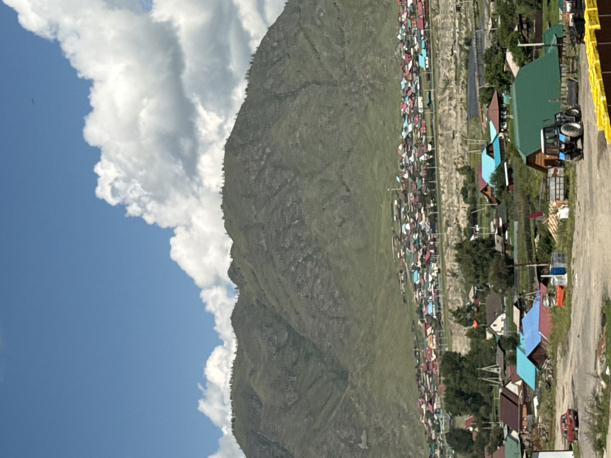The image depicts a rural village nestled at the base of a large, green, and rocky mountain under a bright blue sky with scattered white clouds. The village consists of numerous colorful houses with red, blue, and green roofs. In the foreground, a dirt road is visible, with a red car parked on the left side. A blue tractor is positioned in front of a large green-roofed building. The environment appears to be a remote settlement in a mountainous region, possibly in or around the city of Onguday, Russia, given the architectural style of the houses and the mountainous landscape.