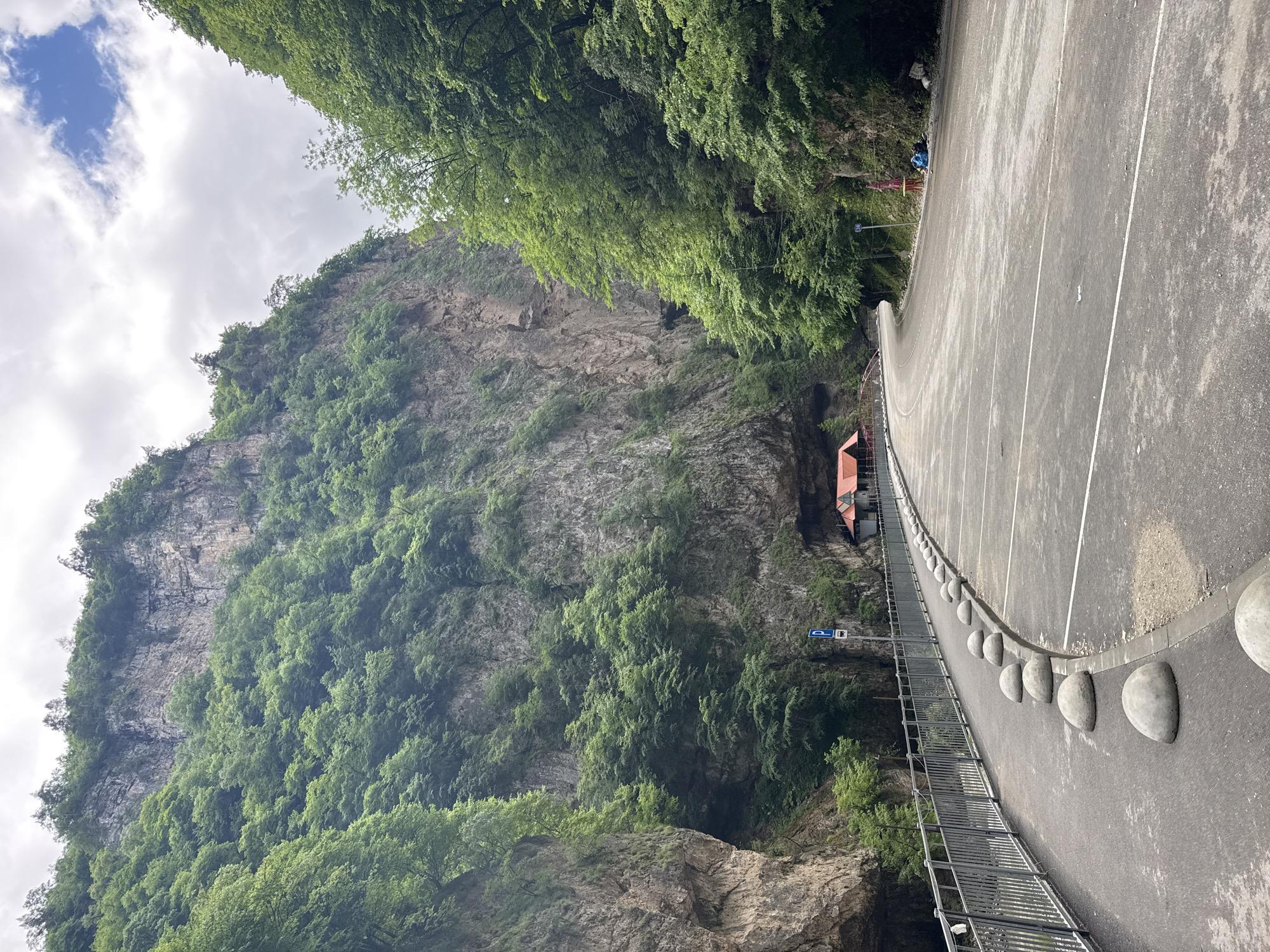 The image shows a scenic view of a mountain gorge with a road winding through it. The gorge walls are steep and covered with lush green trees and vegetation. A paved road with white lane markings curves to the right, bordered by a metal fence and a row of rounded concrete bollards. In the distance, a small red-roofed building is nestled into the side of the gorge, possibly a cafe or rest stop. There are a few people visible near the building and further down the road, some with backpacks. The sky is cloudy with patches of blue. The location is identified as Khushto-Syrt, Russia, a place known for its natural beauty and dramatic landscapes.
