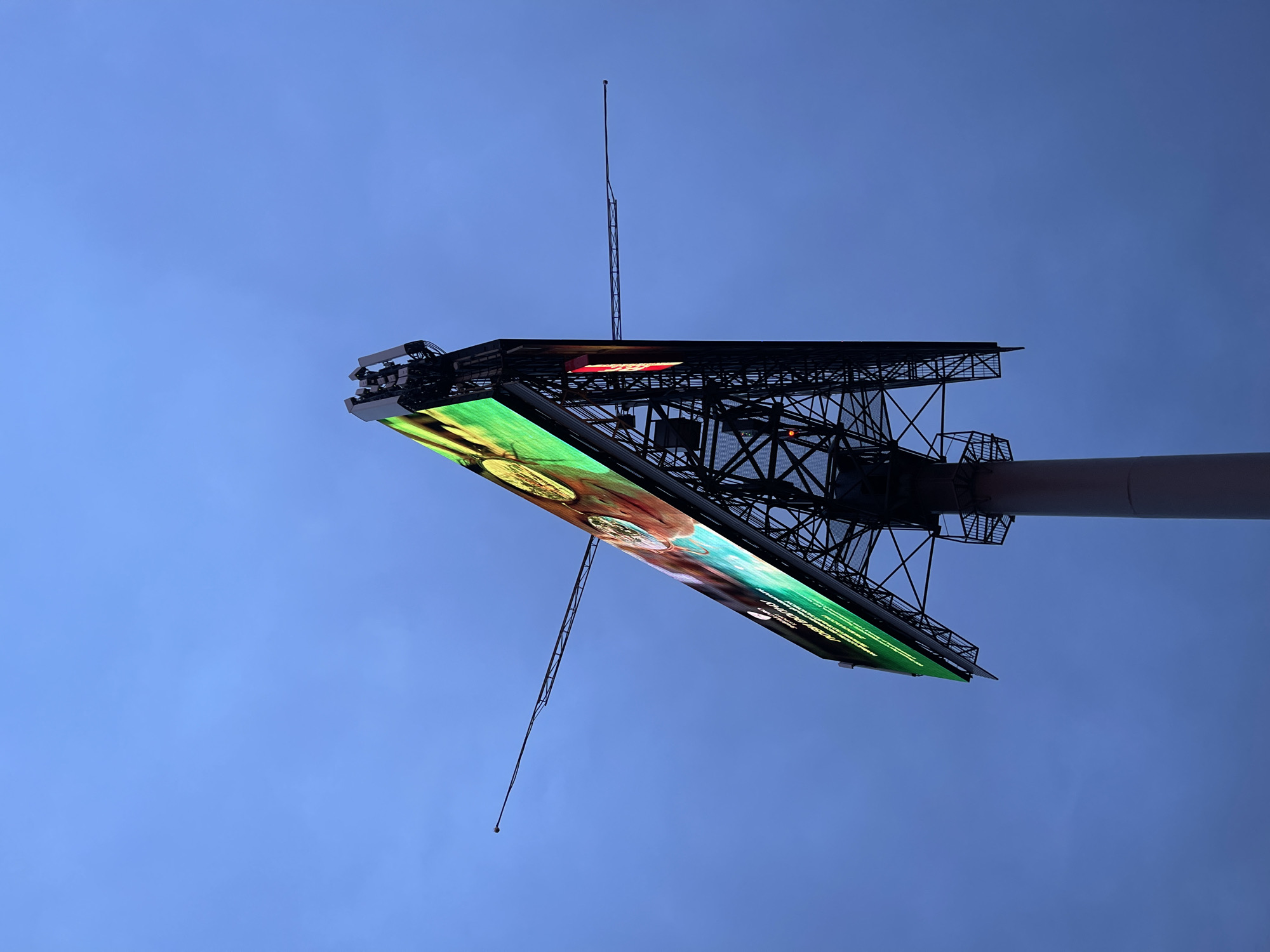 An upward shot showcases a large billboard structure against a solid blue sky in an unknown location. The billboard is supported by a steel framework of beams and braces connected to a central pole. On the visible face of the billboard, bright green and white text is partially visible, with an image of a dish and other text, which are unintelligible. At the top of the billboard's structure are antenna-like rods extending towards the sky.