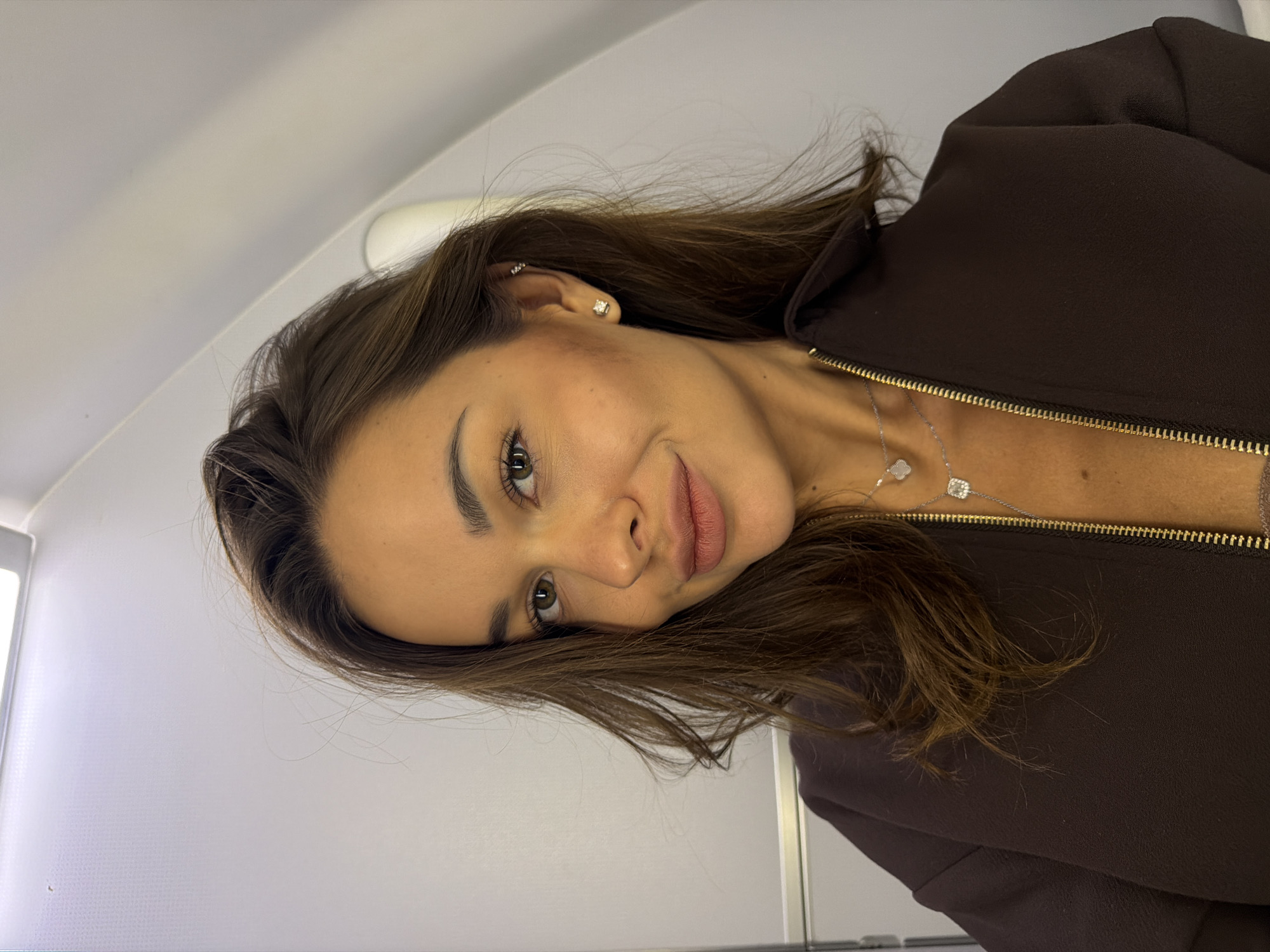 A young woman with brown hair is taking a selfie inside an airplane cabin. She is wearing a dark brown zippered jacket, a nude-colored top, and two silver necklaces. Her left ear has two piercings, with diamond stud earrings in both her upper and lower earlobe. She is smiling faintly, looking directly at the camera. The background shows the interior of an airplane, with white walls and a ceiling light. The location is within the city of Перепечино, Russia.