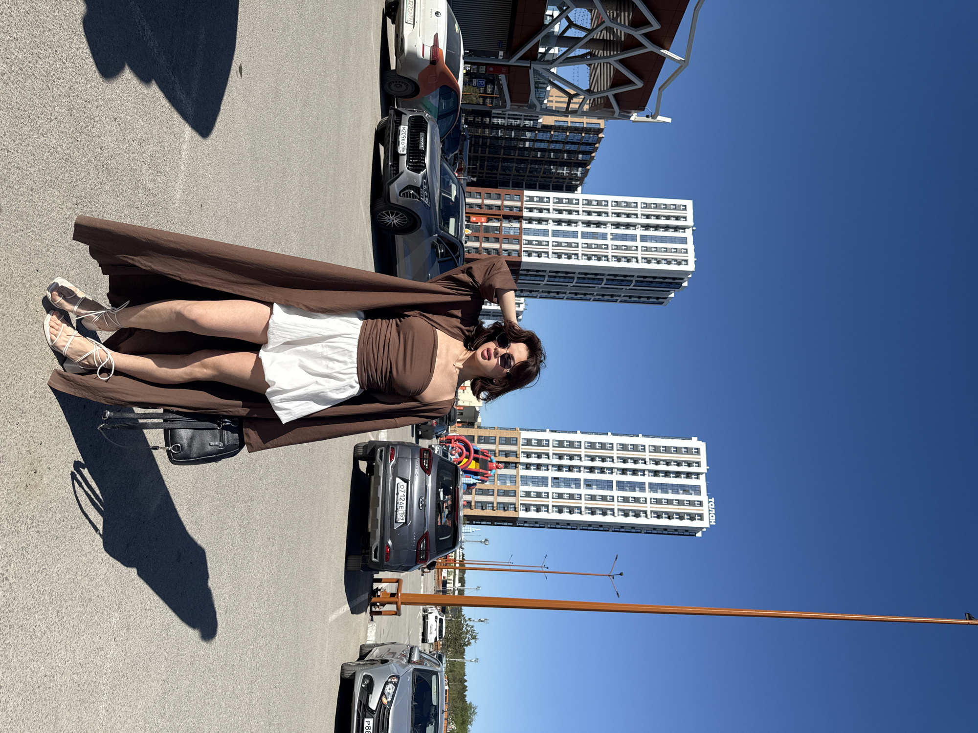 A woman stands in a parking lot in Perm, Russia on a sunny day. She wears a brown top and a white skirt, covered by a long brown coat. She is also wearing sunglasses and strappy sandals. She stands next to a black purse. The parking lot has several cars. In the background are tall, white high-rise buildings and a blue sky.