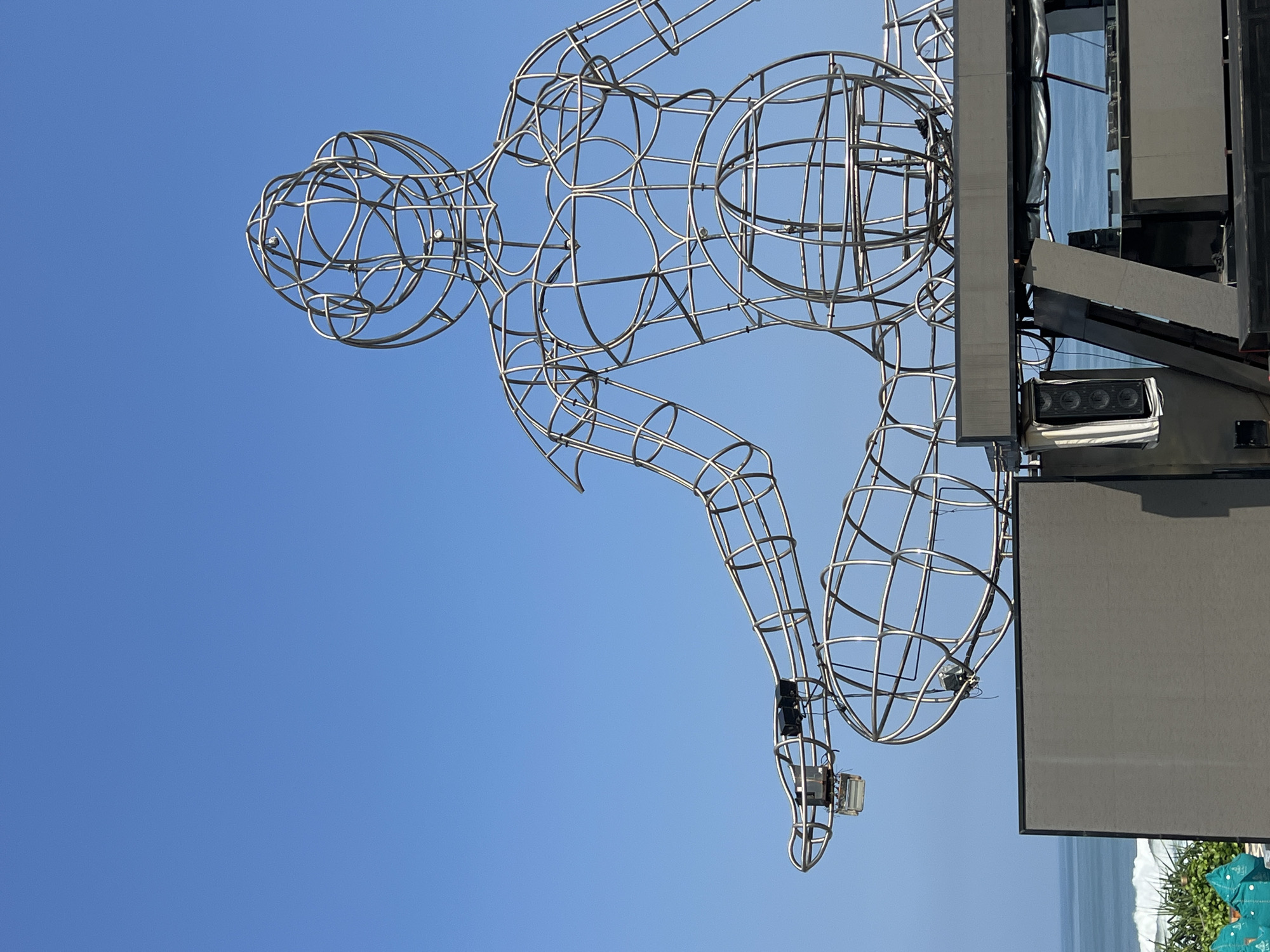 The image captures a large, metallic wireframe sculpture of a seated figure, resembling a meditative pose, against a clear blue sky. The sculpture is intricate, with its skeletal structure composed of curved metal rods forming the head, torso, arms, and legs. The figure appears to be positioned on top of a dark, multi-level stage structure, which includes large screens and speakers. In the background, a body of water, likely the ocean, is visible with gentle waves breaking near the shore. Green foliage can be seen in the lower left corner. The lighting suggests it is daytime. The sculpture is located at the Garuda Wisnu Kencana Cultural Park in Ungasan, Badung, Bali, Indonesia.