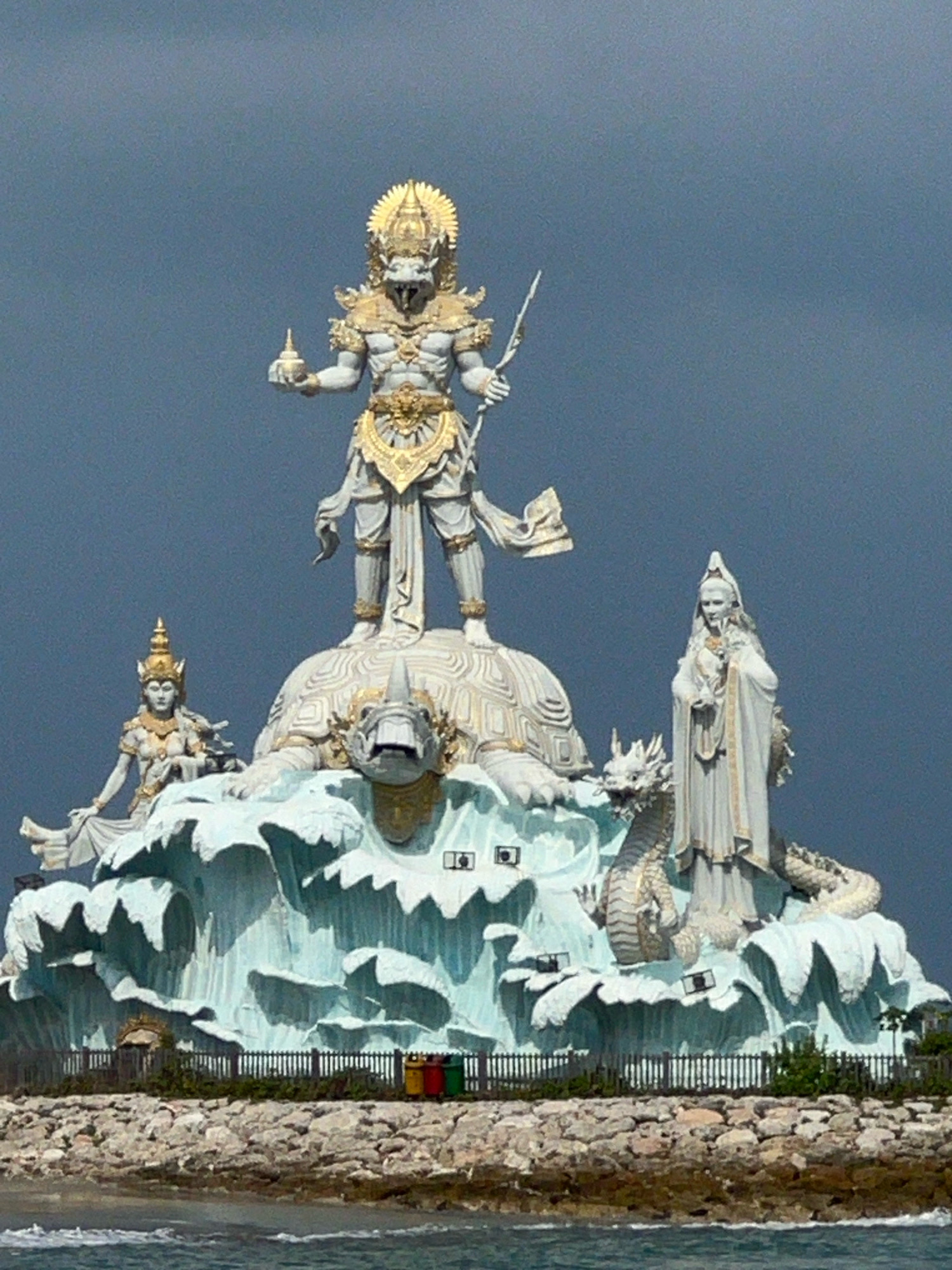 The image depicts the Garuda Wisnu Kencana (GWK) Cultural Park in Kuta, Indonesia. The primary subject is a colossal statue of Vishnu riding on the back of Garuda, an eagle-like creature. The statue is predominantly white with gold accents. Vishnu, depicted with a reptilian head and muscular build, stands tall, holding a spear in his right hand and a vessel in his left. Below Vishnu, Garuda is represented as a turtle, with its head visible between Vishnu's legs. To the left of Vishnu is a statue of a seated female figure adorned with a crown and jewelry. To the right of Vishnu stands another female figure in flowing robes, holding what appears to be a flower. The statues are positioned on a large, stylized wave-like structure, which is light blue and white. A dragon statue is also incorporated into the base, with its body wrapping around the figures. In the foreground, there is a rocky shoreline with a wooden fence and several trash bins (yellow, red, and green). The sea is visible at the bottom of the frame. The sky above is a dark, overcast gray, suggesting a cloudy or stormy day.